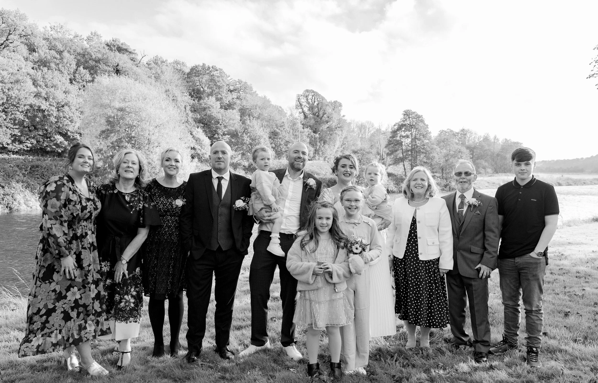 A group of fifteen people, including children, standing outdoors on grass near a river, smiling for a photo with trees and water in the background.