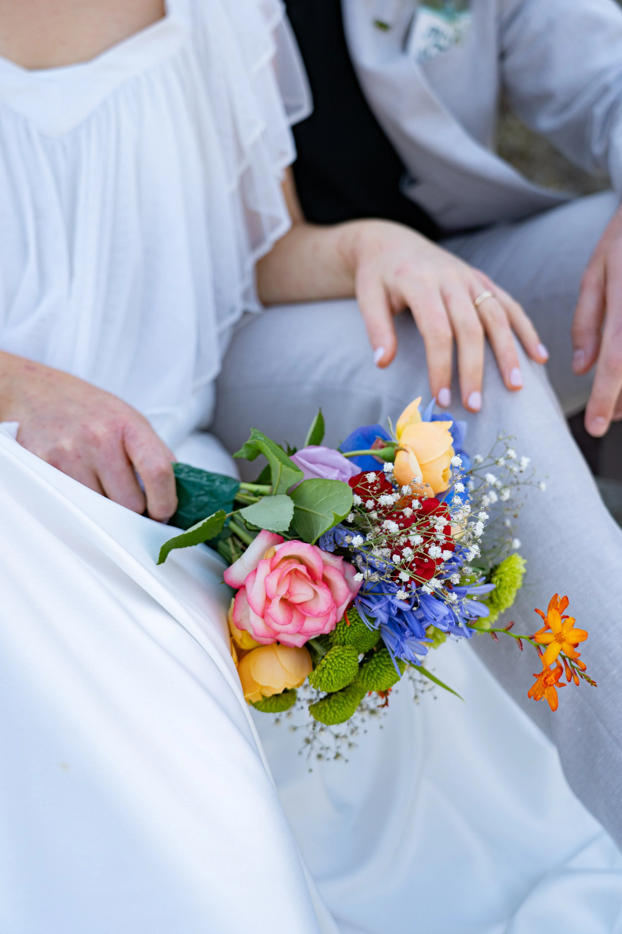 Close-up of a person holding a colorful bouquet of flowers resting on their lap, with another person sitting nearby.