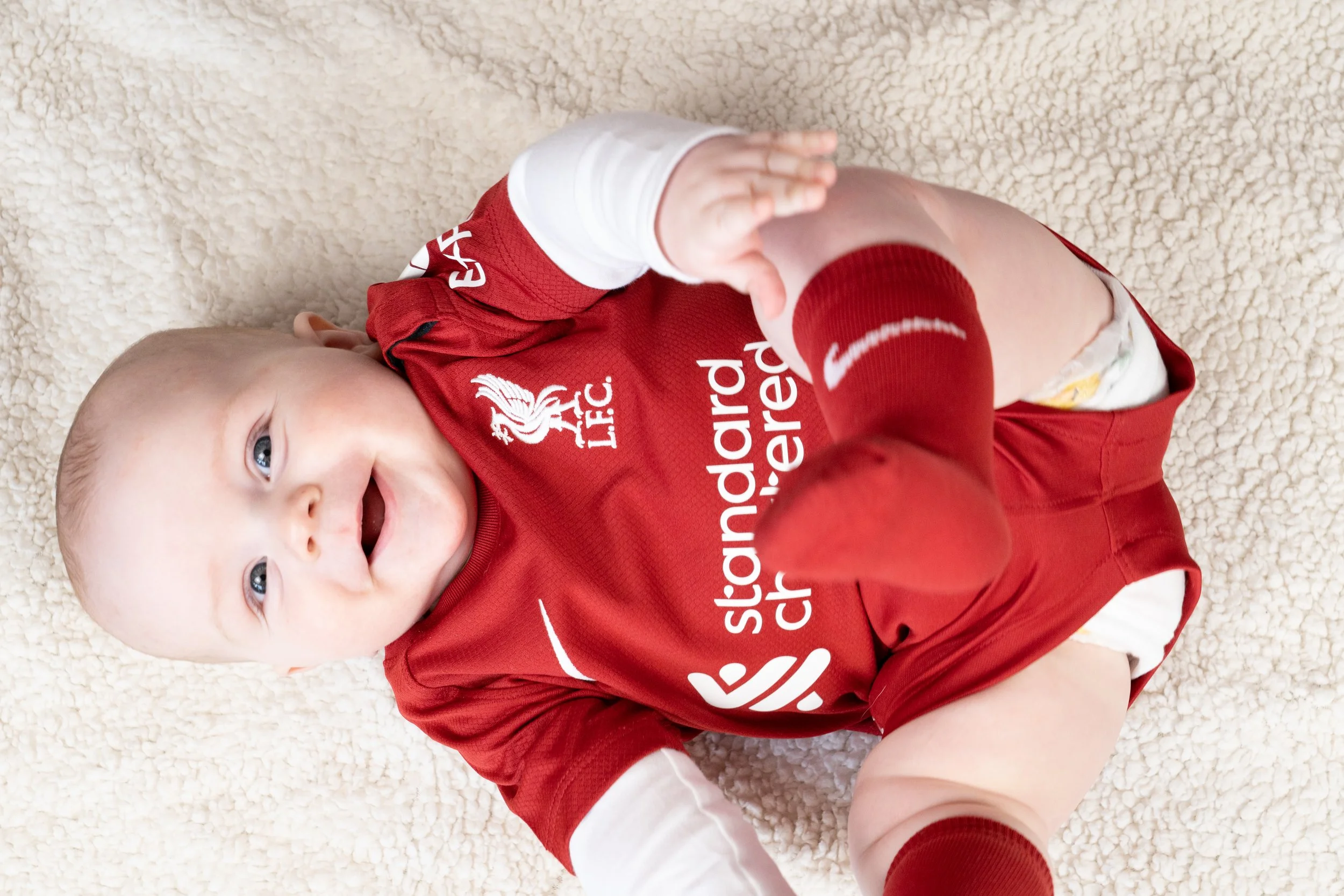 Infant lying on a soft white blanket, smiling, wearing a red Liverpool FC jersey and red socks.