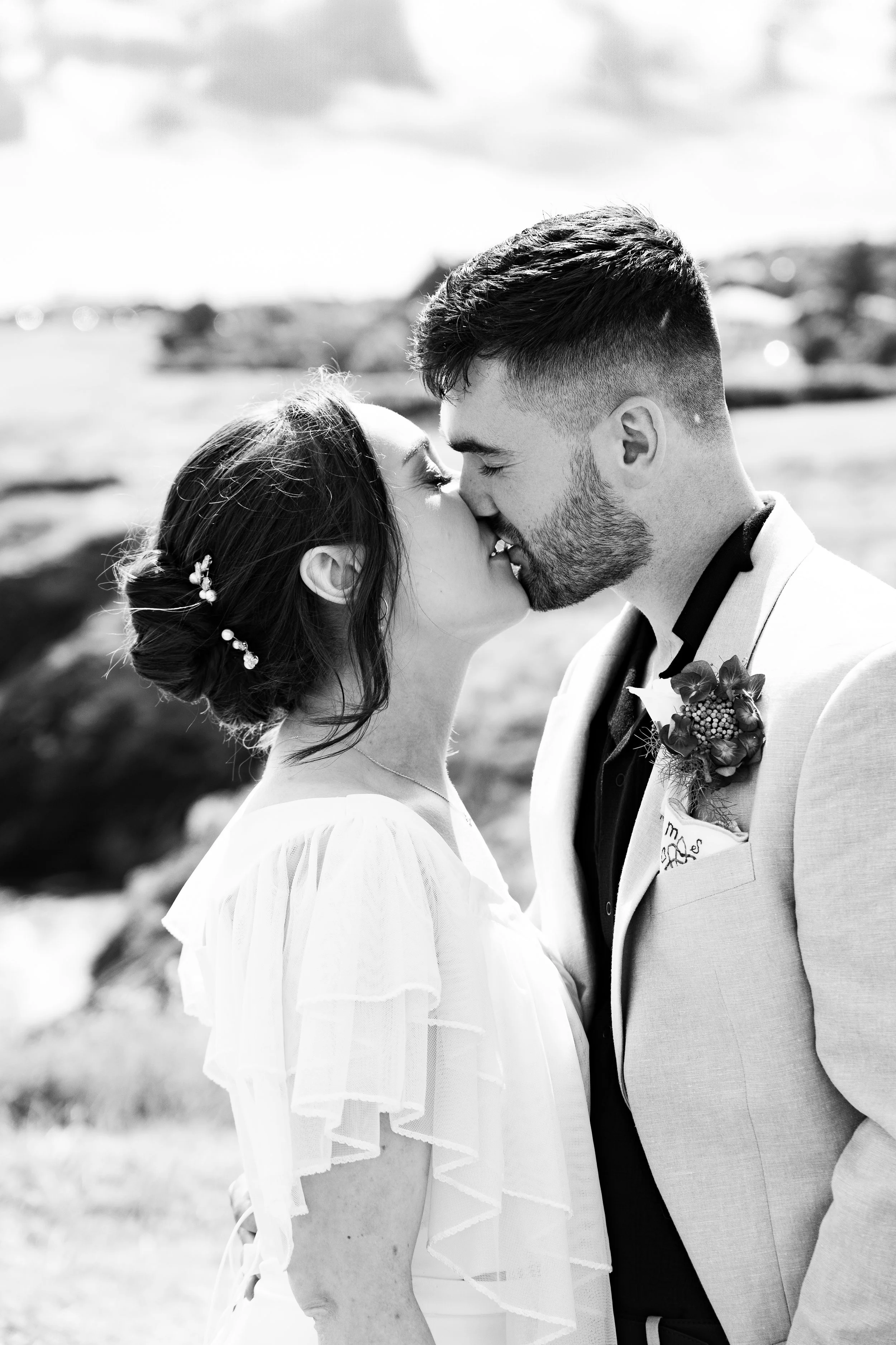 A black and white photo of a bride and groom kissing outdoors, with a rocky landscape and water in the background.