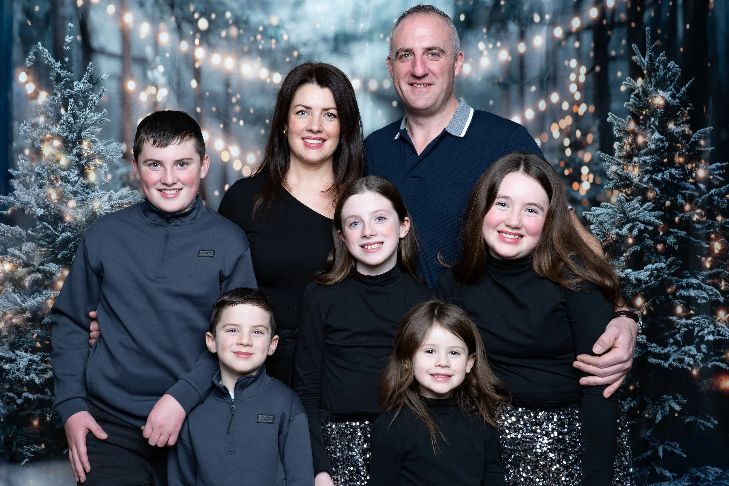 Family portrait of a man, a woman, and five children standing in front of a snowy, Christmas-themed backdrop with decorated trees and string lights.