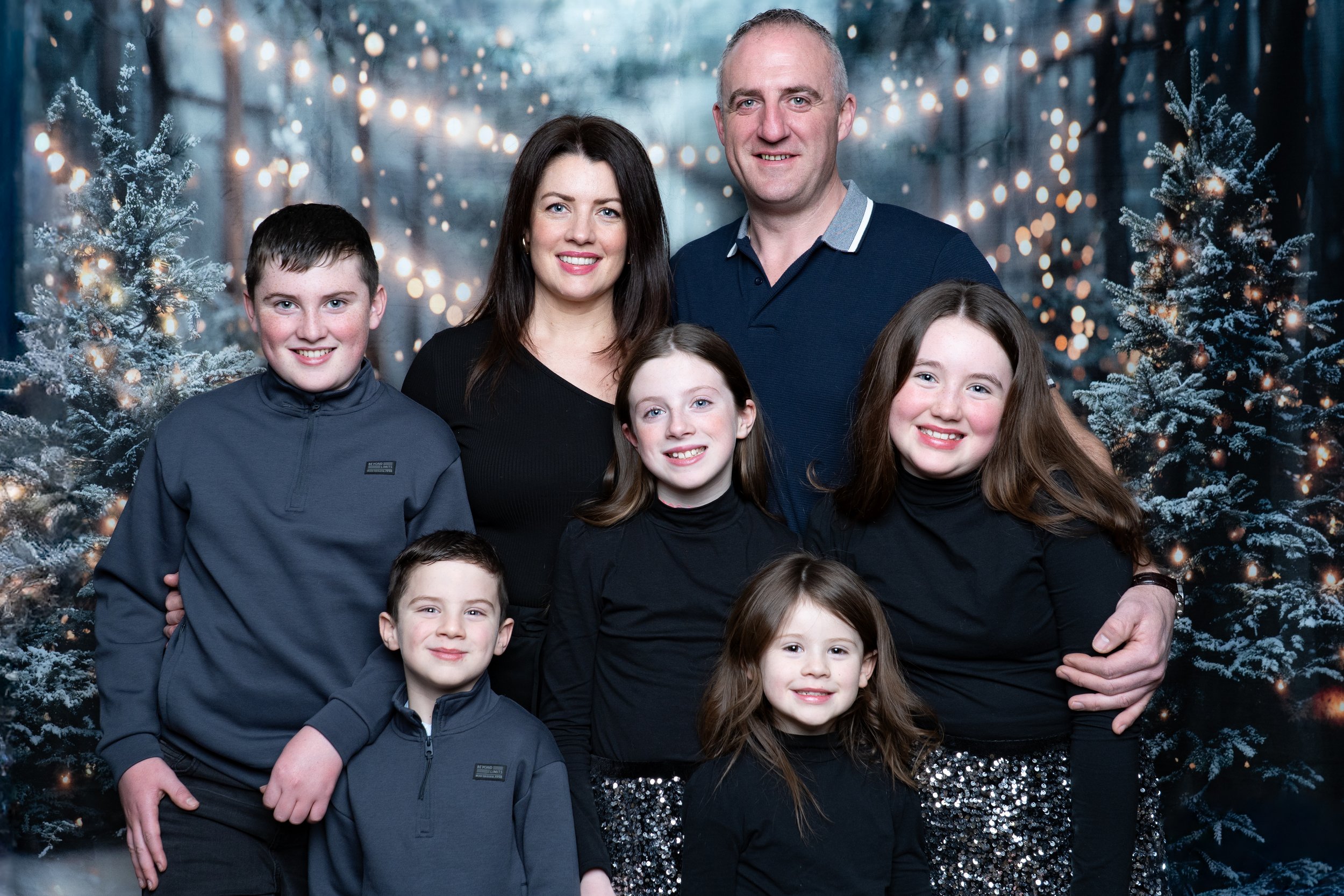 Family portrait of a man, a woman, and five children standing in front of a snowy, Christmas-themed backdrop with decorated trees and string lights.
