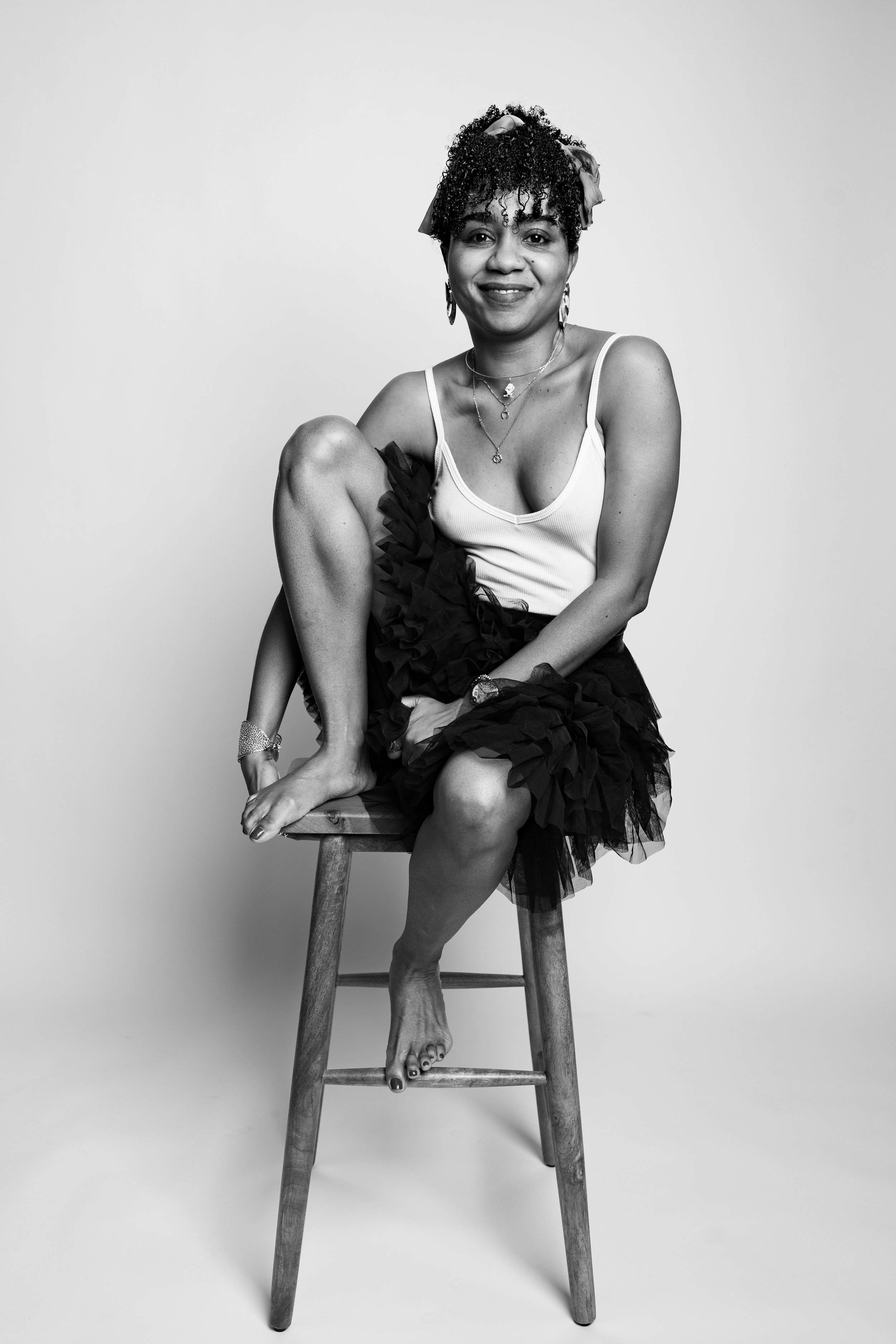 Black and white portrait of a woman sitting on a wooden chair, smiling, with one knee pulled up to her chest and the other foot on the ground. She has curly hair, wears a sleeveless top, a ruffled skirt, and jewelry, set against a plain light backgro