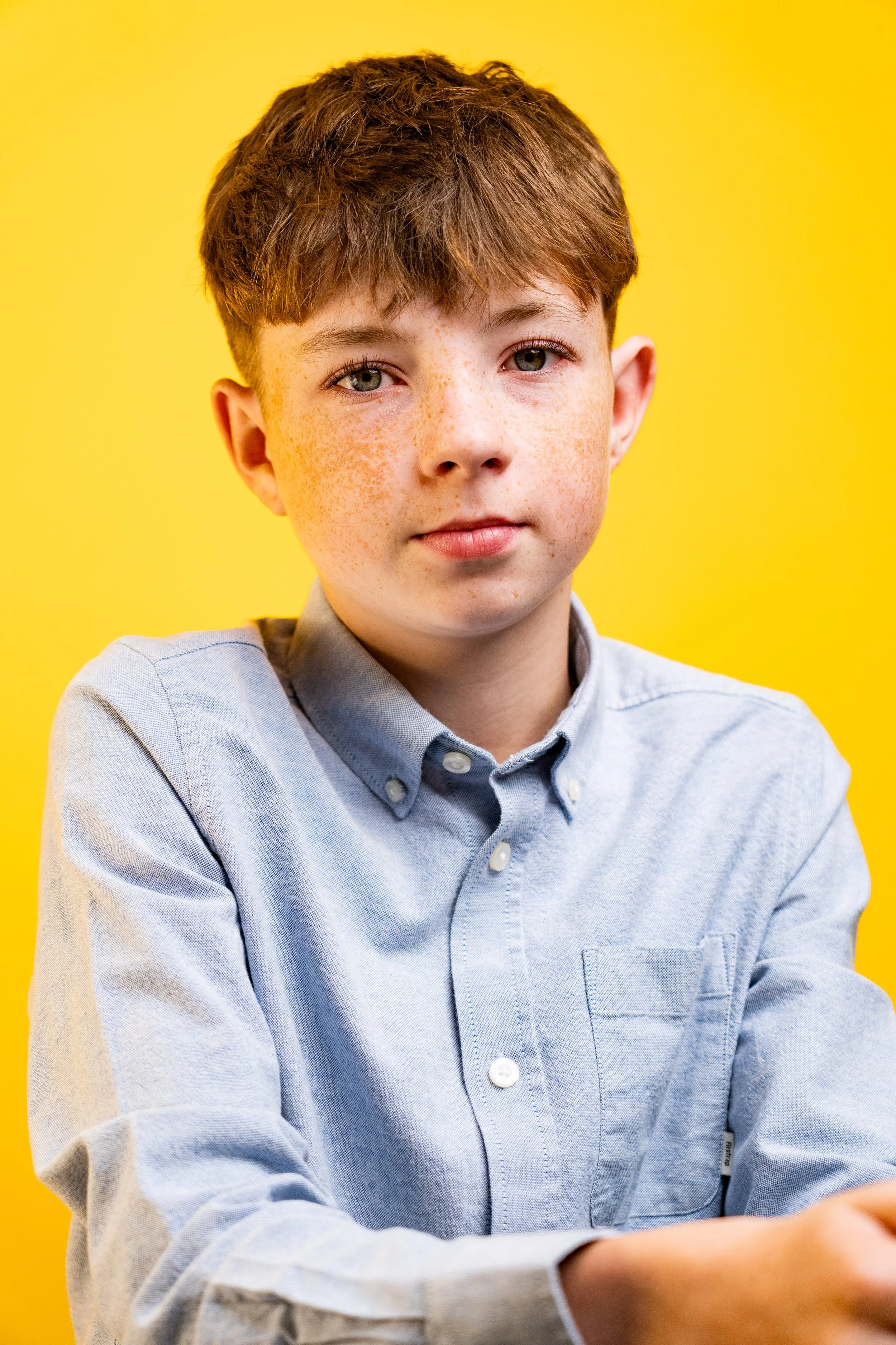 Portrait of a young boy with brown hair and freckles, wearing a light blue button-up shirt, against a yellow background.