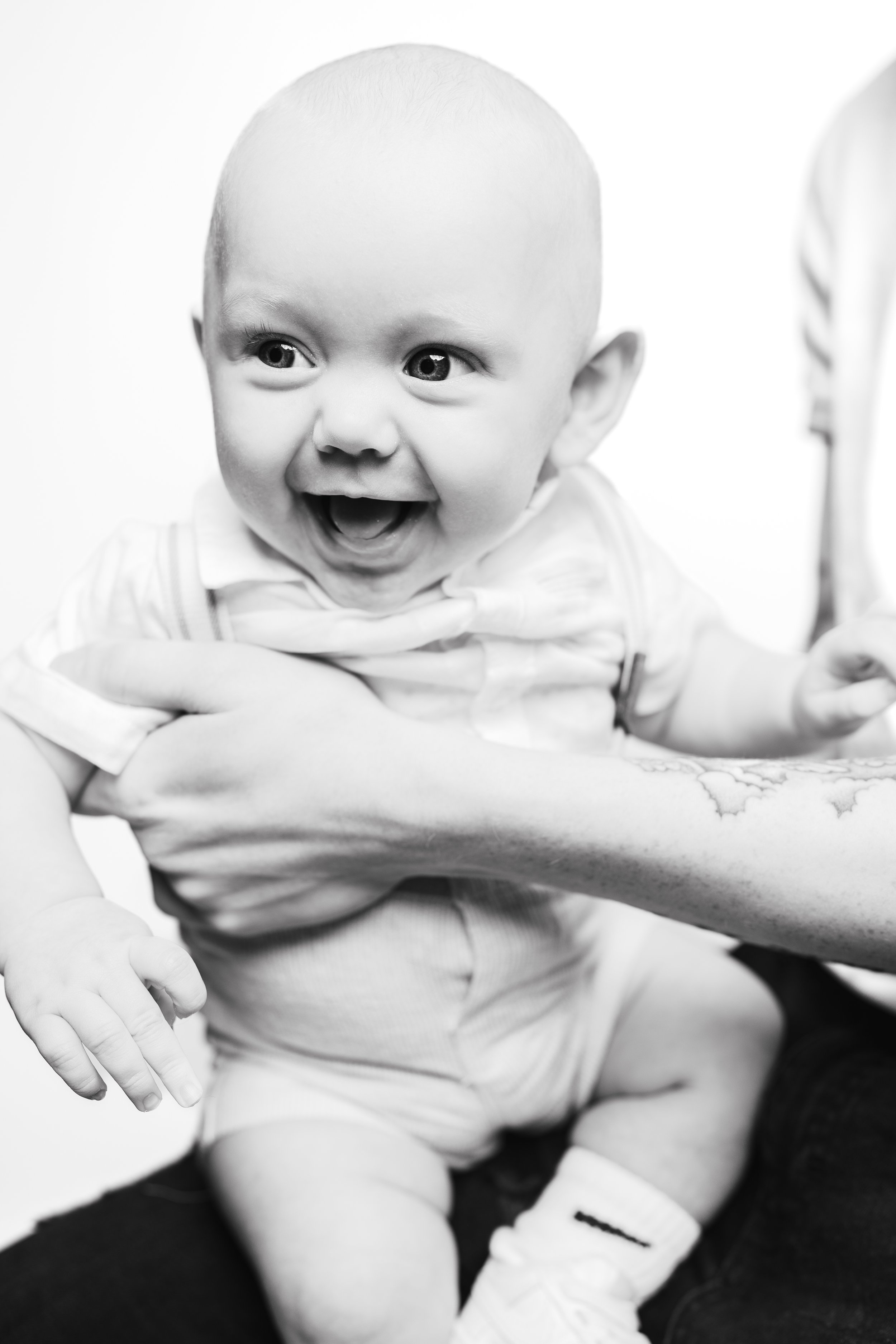A happy baby with light skin, big eyes, and a wide open mouth, being held by an adult with tattooed arms, in black and white.