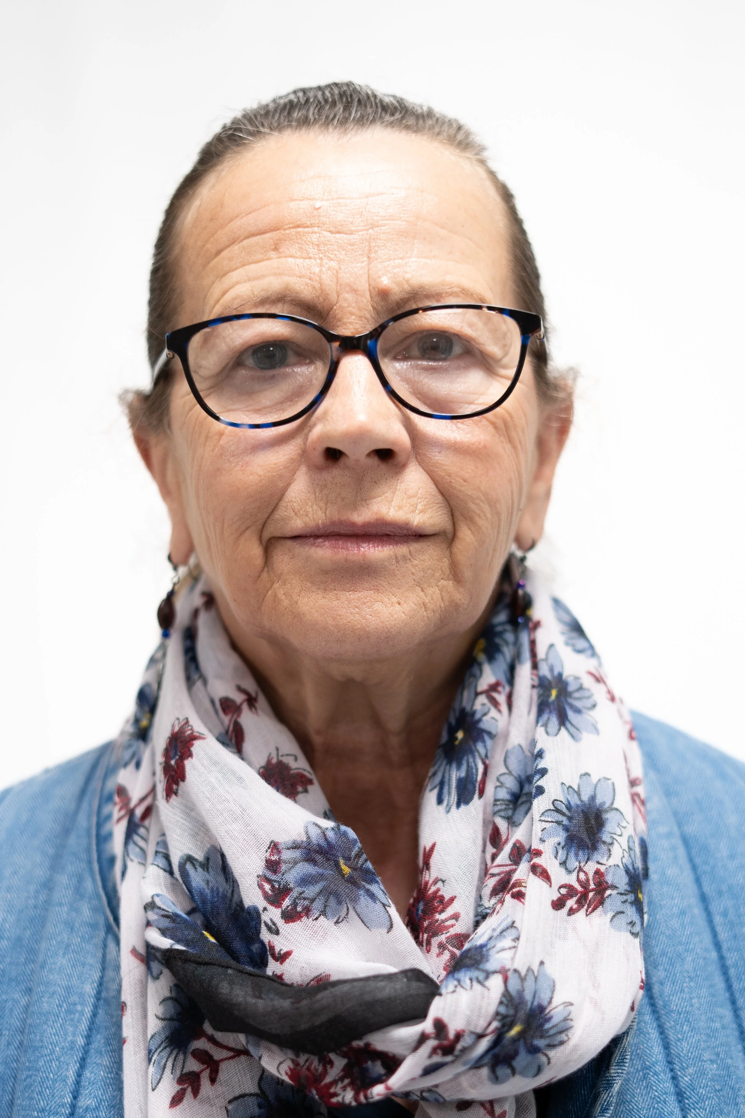 Close-up portrait of an elderly woman with glasses, wearing a floral scarf and blue cardigan, against a plain white background.