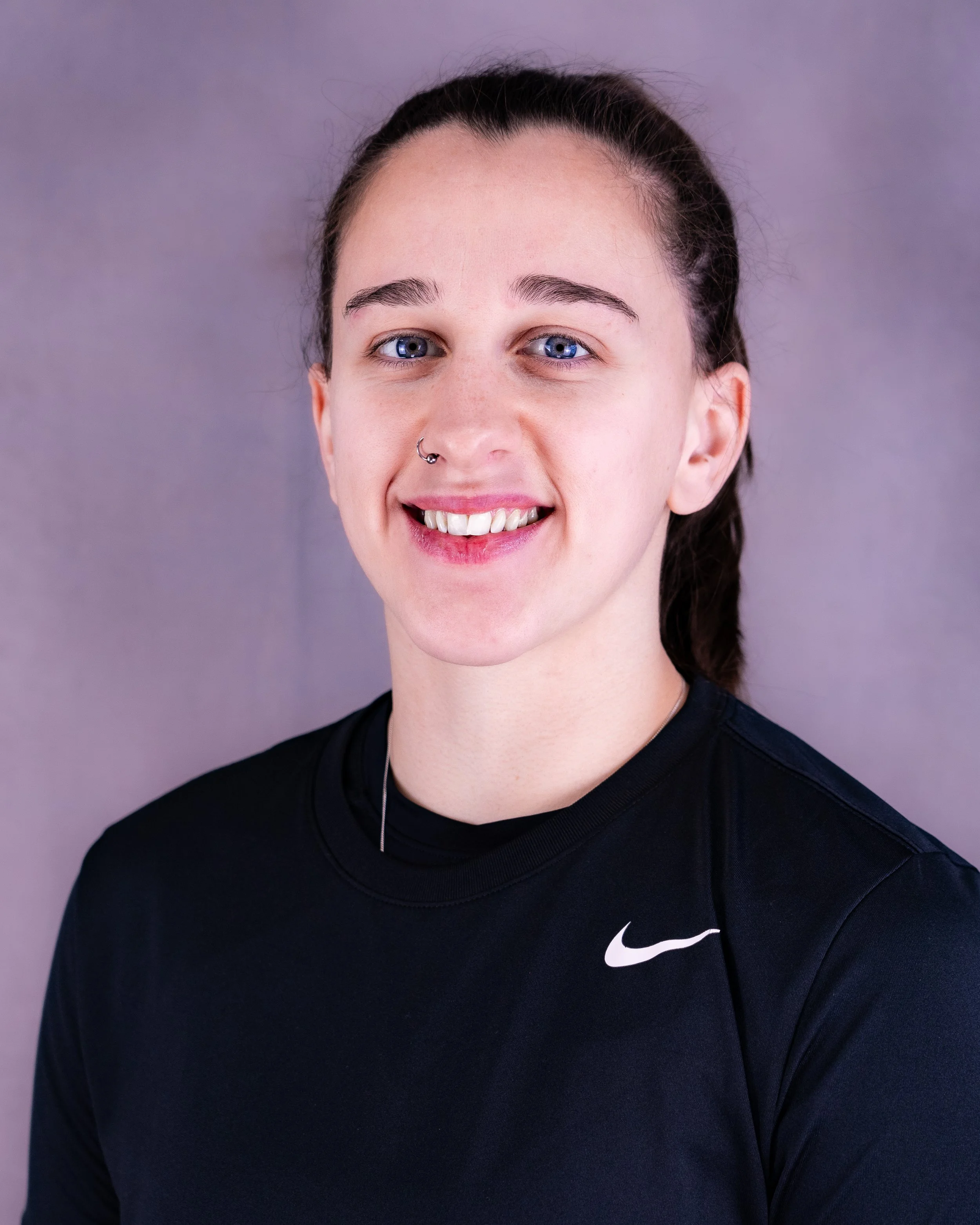 A young woman with fair skin, blue eyes, and dark hair in a ponytail, smiling and wearing a black Nike athletic shirt, against a purple background.