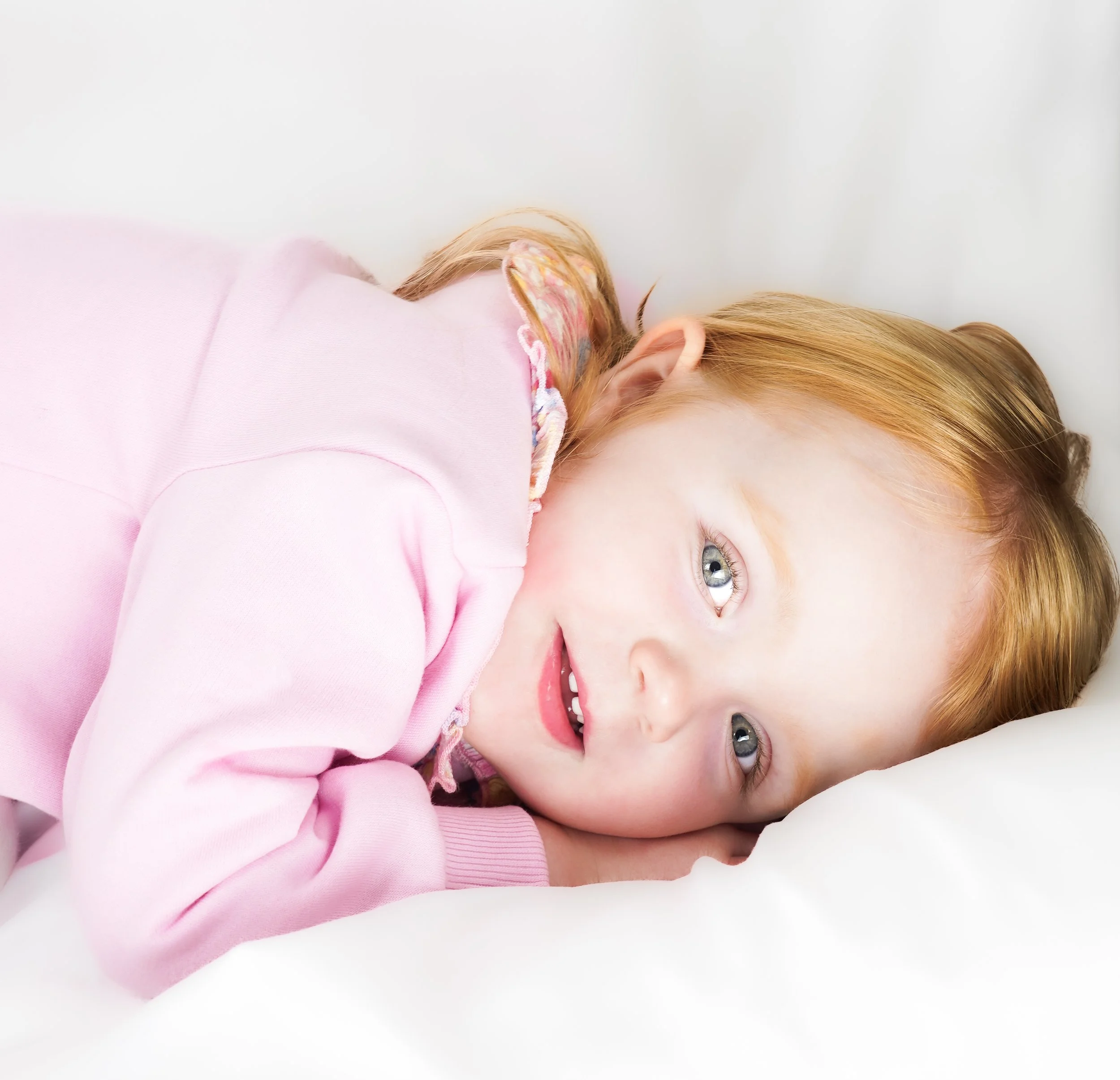 A young girl with red hair and blue eyes lying on her side on a white pillow, wearing a pink long-sleeve shirt, smiling and looking happy.
