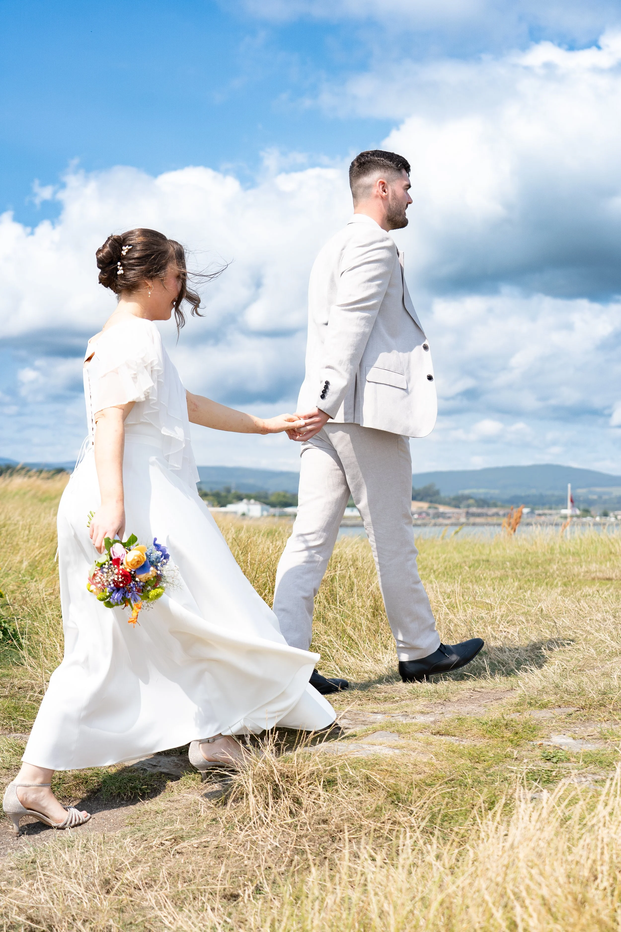 A bride and groom walking outdoors in a field, holding hands. The bride is wearing a white wedding dress and carrying a colorful bouquet. The groom is dressed in a light-colored suit, and the sky is partly cloudy.