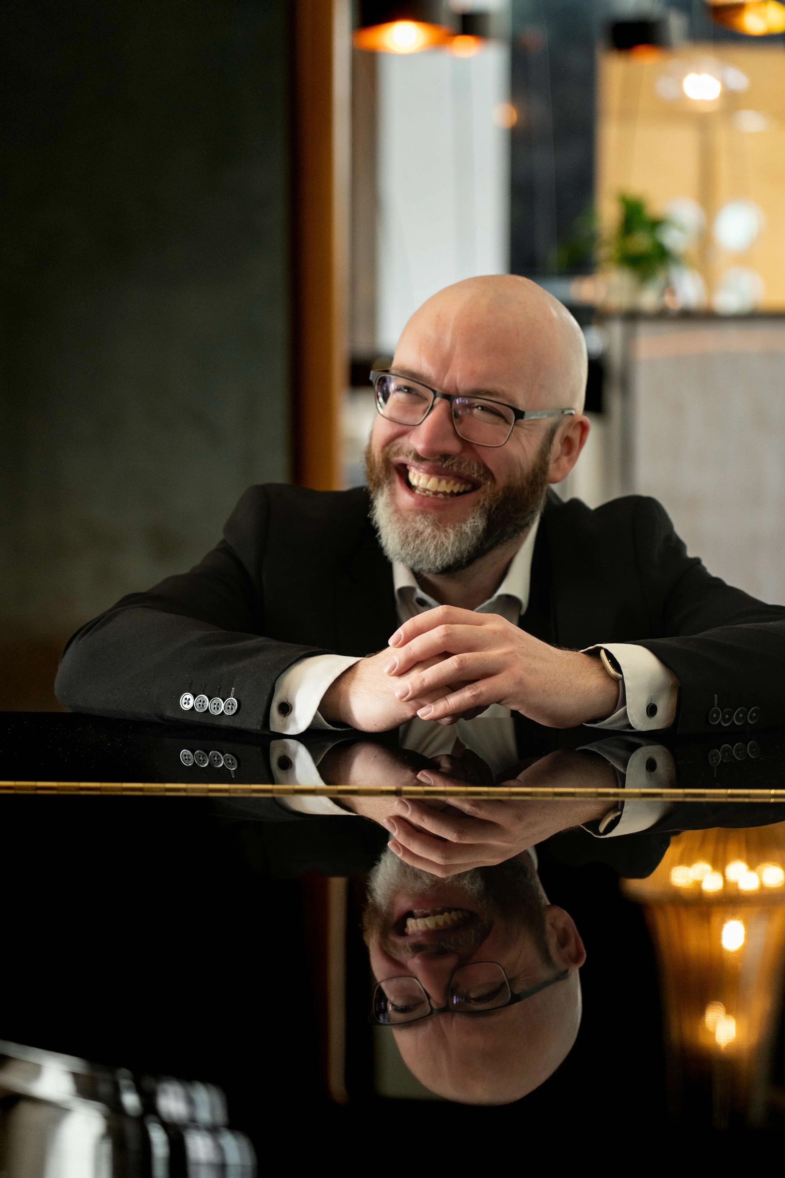 A man with glasses, a beard, and a bald head is smiling and sitting at a reflective black surface, possibly a piano, in an indoor setting with warm lighting.