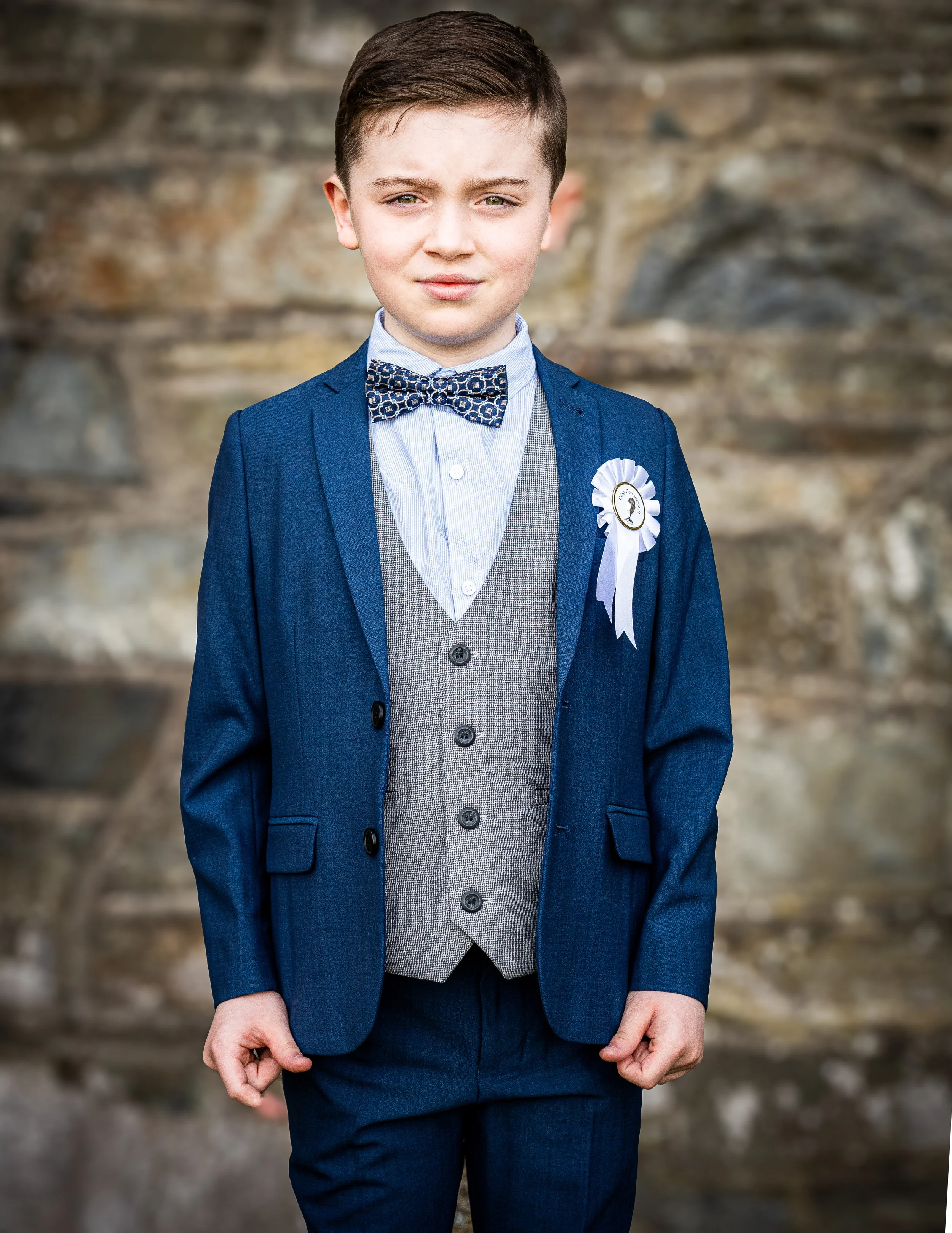 A young boy in a blue suit, gray vest, white shirt, and patterned bow tie standing outdoors in front of a stone wall, wearing a white ribbon badge.