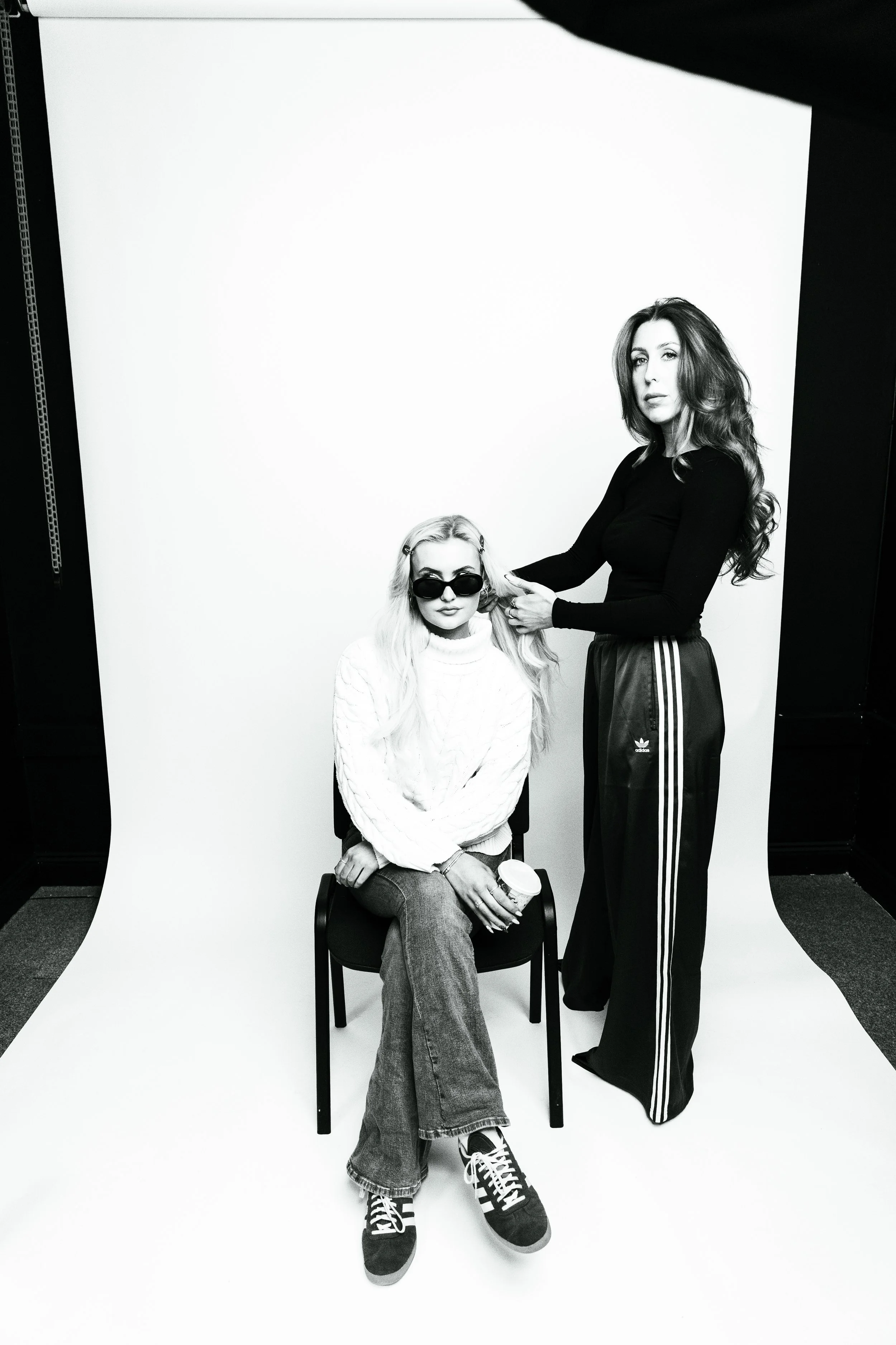 Black and white photo of two women in a studio with a plain white background. One woman, sitting on a chair, has long, blonde hair, sunglasses, a white sweater, jeans, and sneakers, holding a coffee cup. The other woman, standing beside her, has long, wavy hair, and wears a black long-sleeve top and track pants.