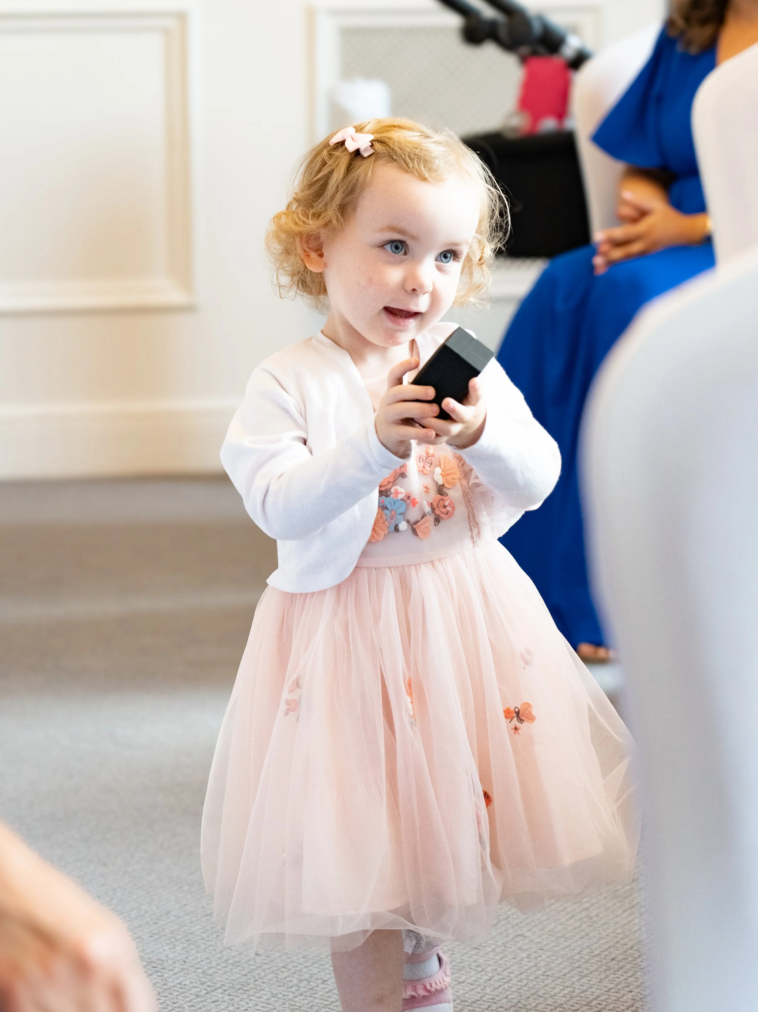 A young girl with curly blonde hair and a pink bow, wearing a pink tulle dress with floral embellishments and a white cardigan, holding a black remote control.