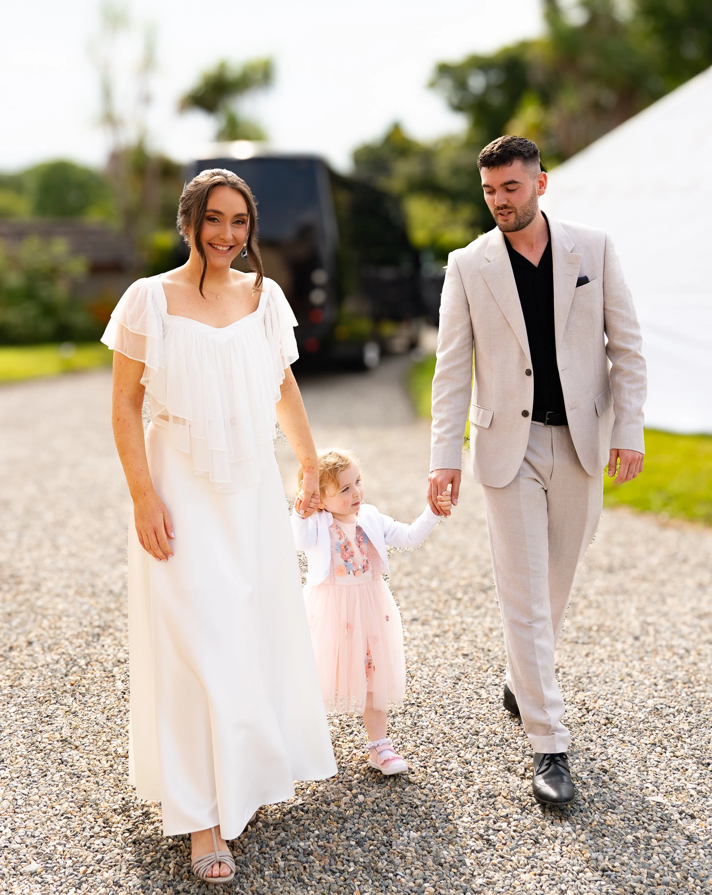 A woman in a white dress and a man in a beige suit hold hands with a young girl in a pink dress, walking outdoors at a gathering or celebration.