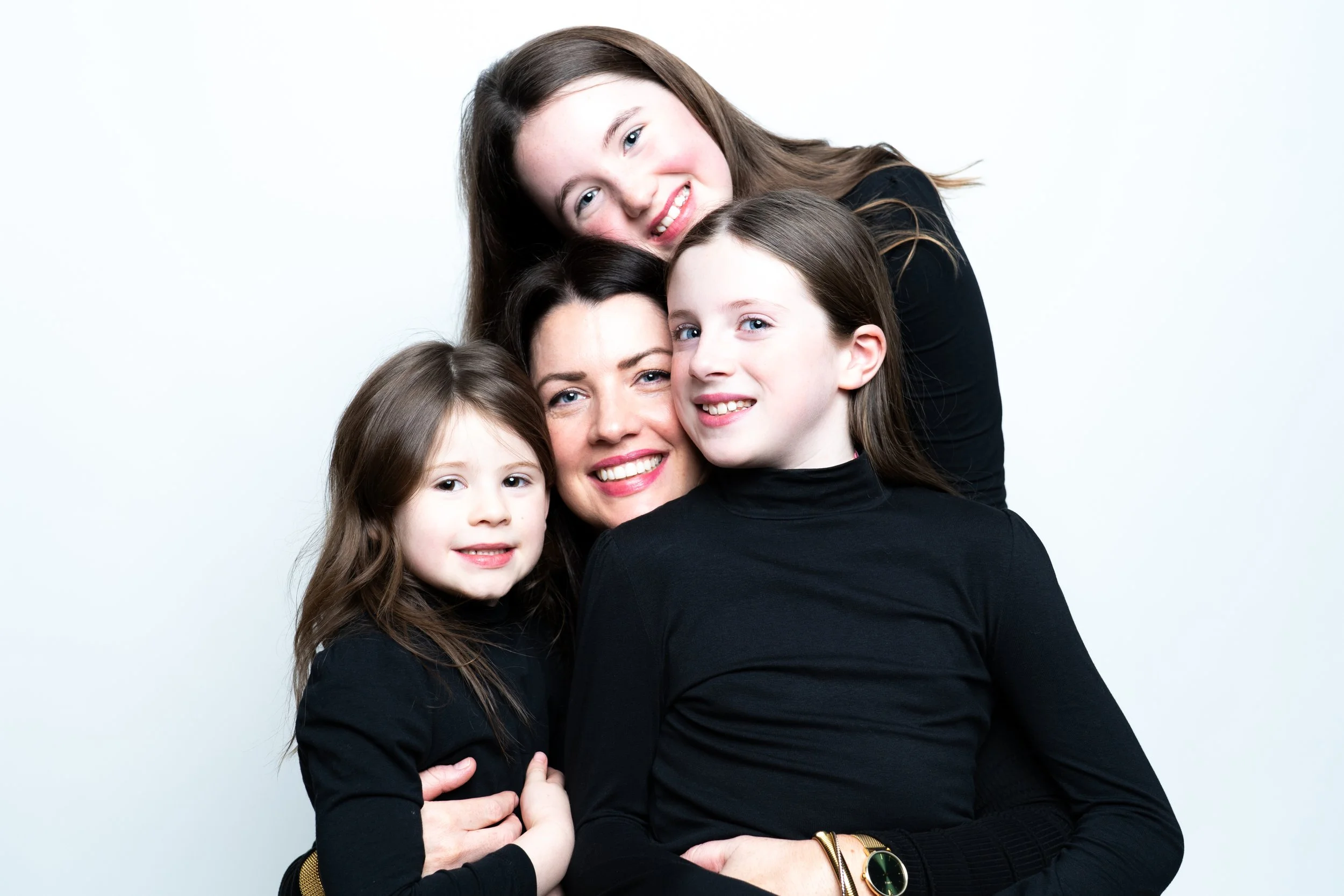 Four women, including an adult, smiling and posing together in black tops against a white background.
