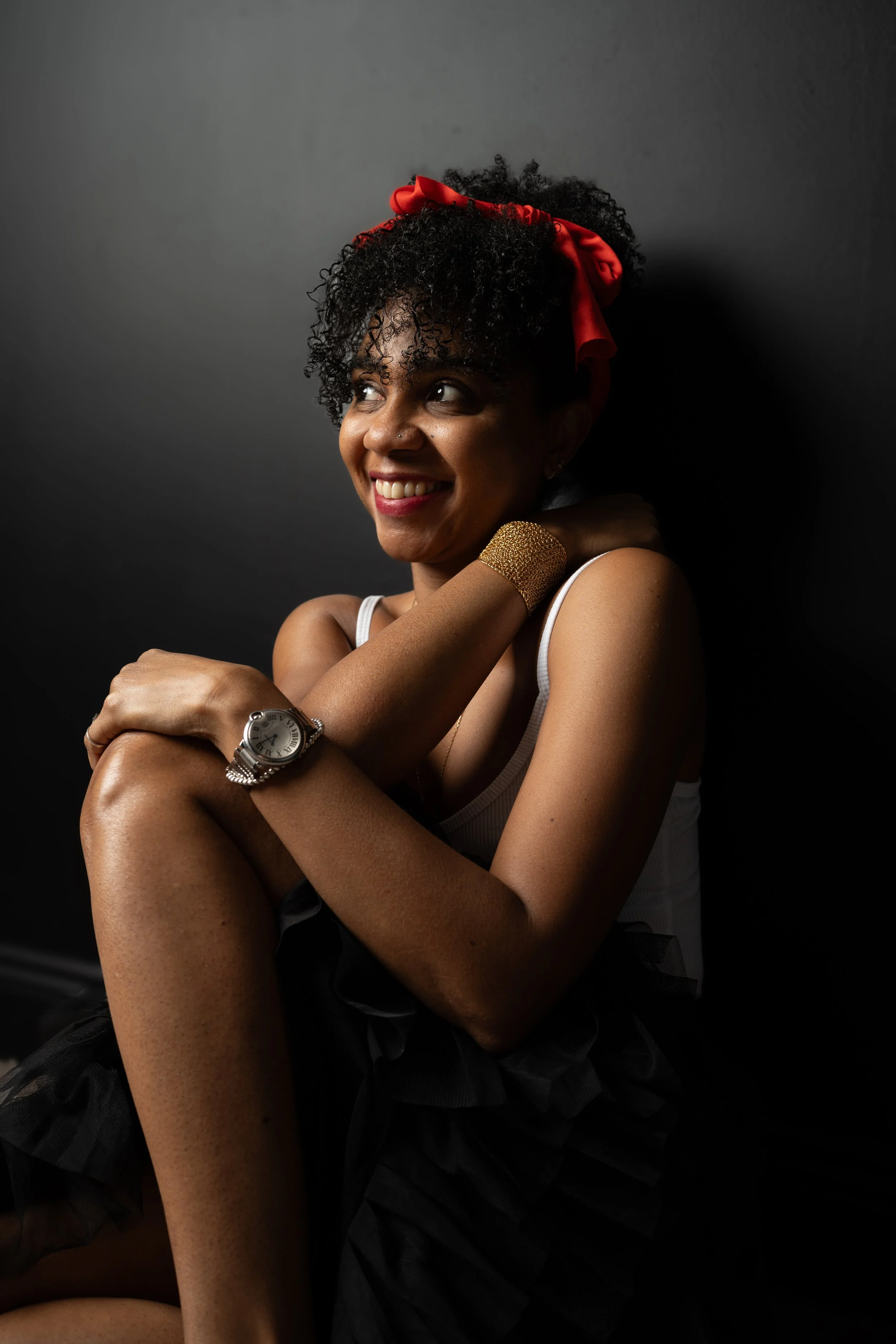 A woman with curly hair wearing a red headband, gold jewelry, and a watch, smiling and sitting against a dark background.