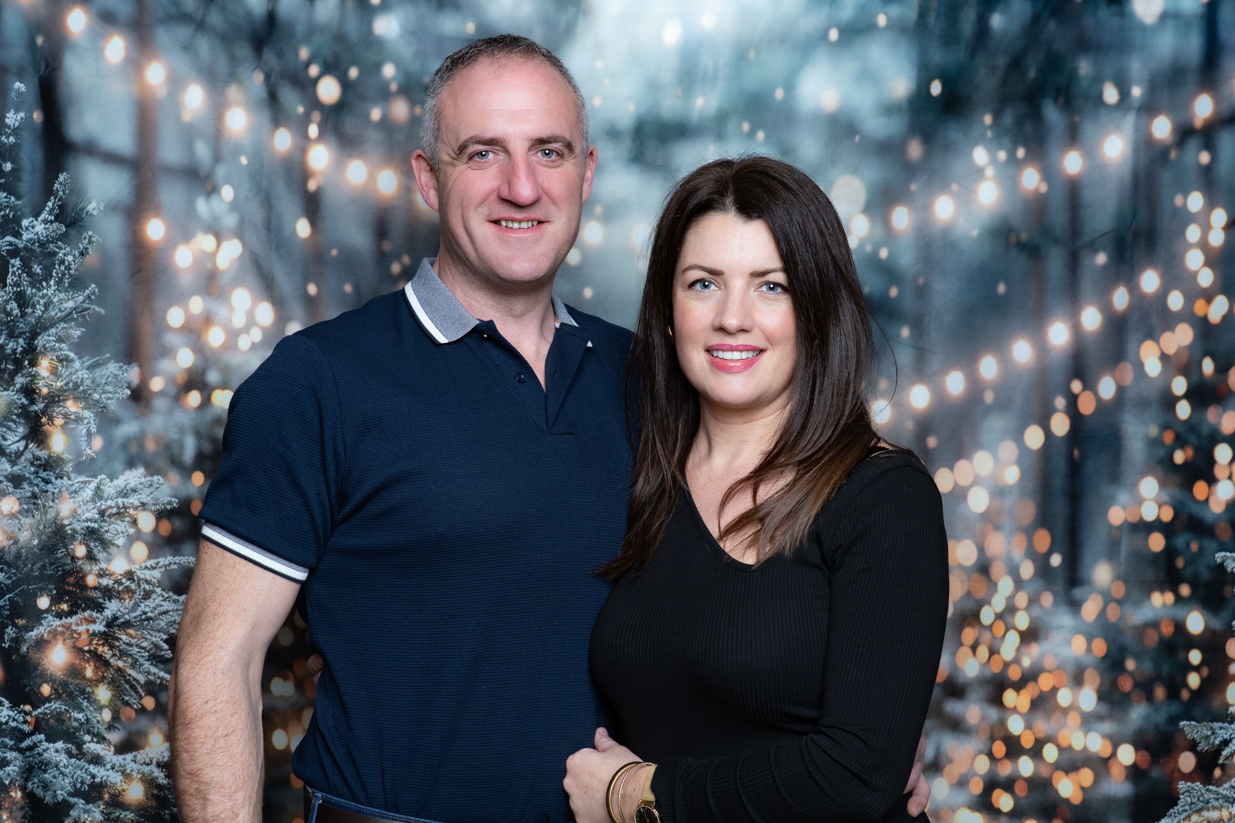 A smiling couple posing together in front of a festive, blurred outdoor background with Christmas trees and string lights, dressed casually.