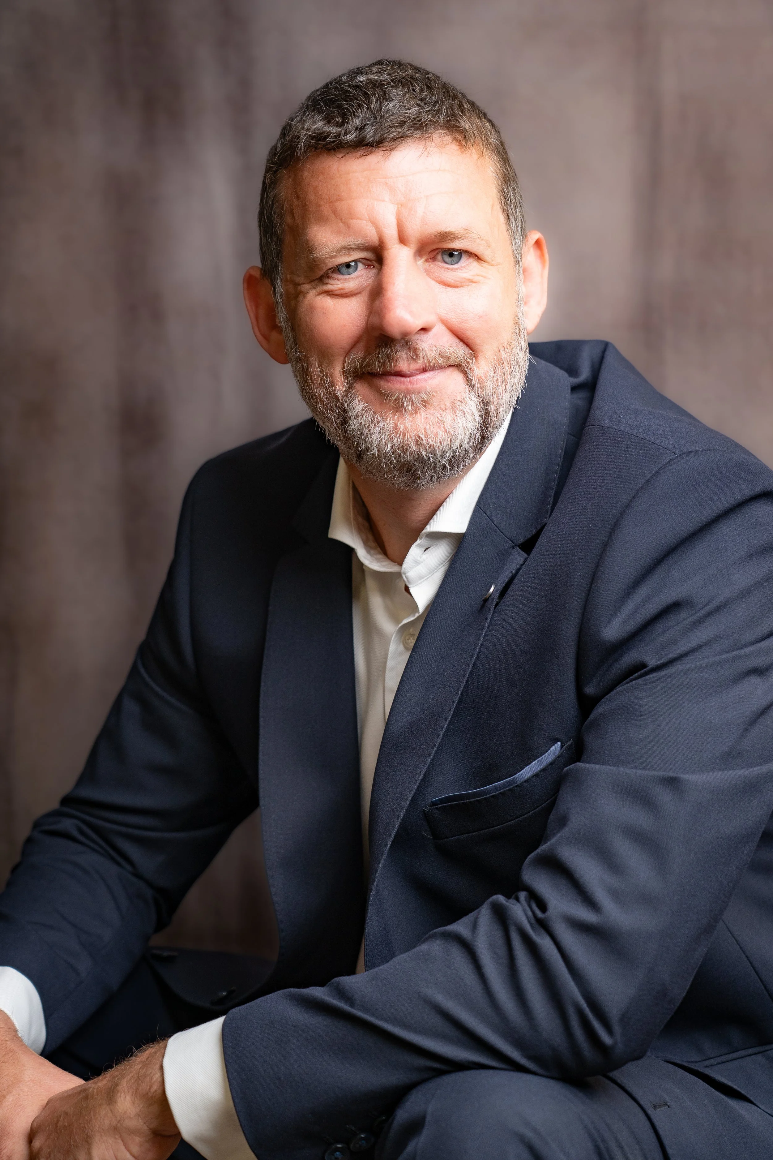 A portrait of a middle-aged man with a beard and short hair, wearing a navy blue suit and white shirt, sitting against a brown background.