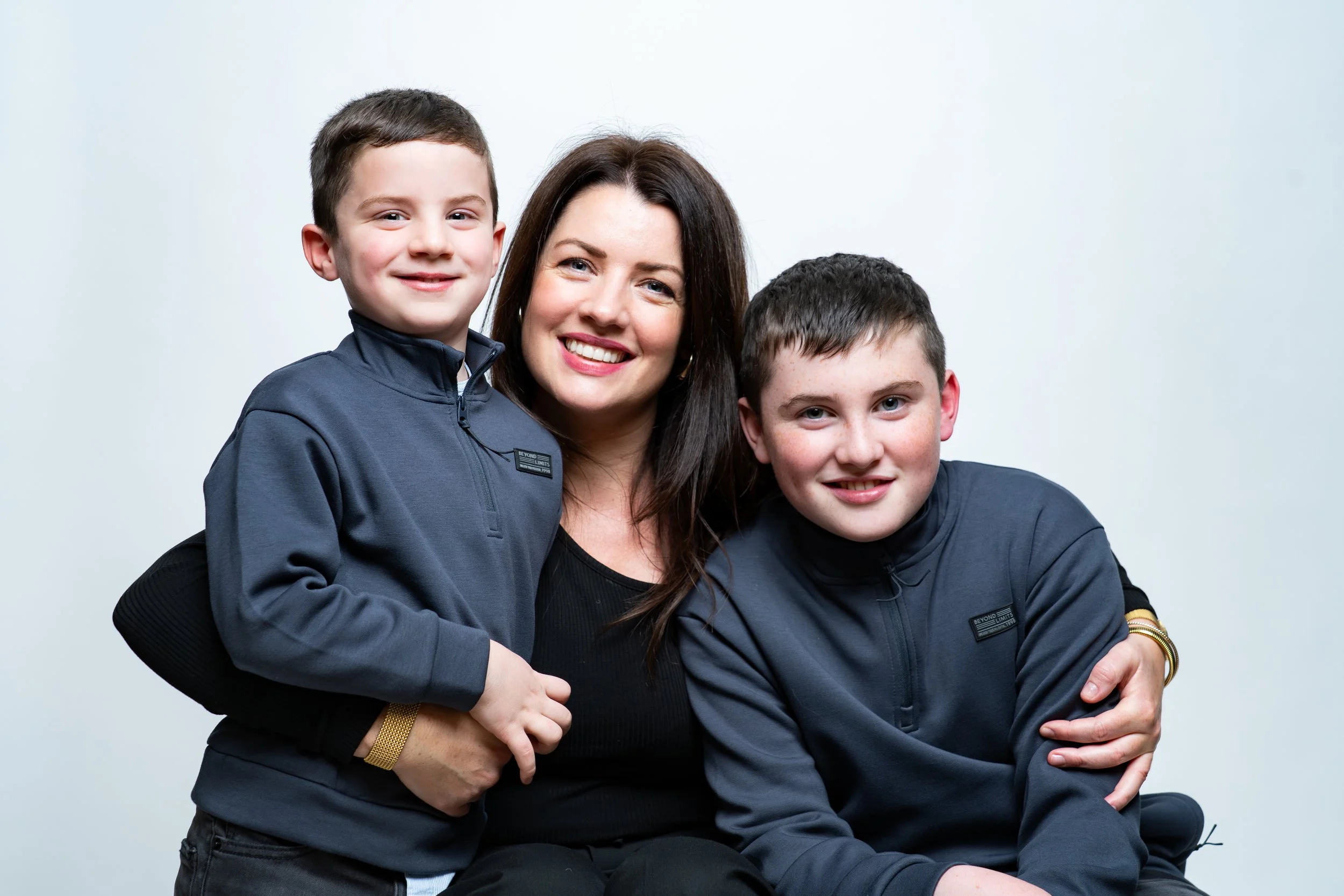 A woman with two young boys, all smiling and wearing dark clothing, posing together against a plain white background.