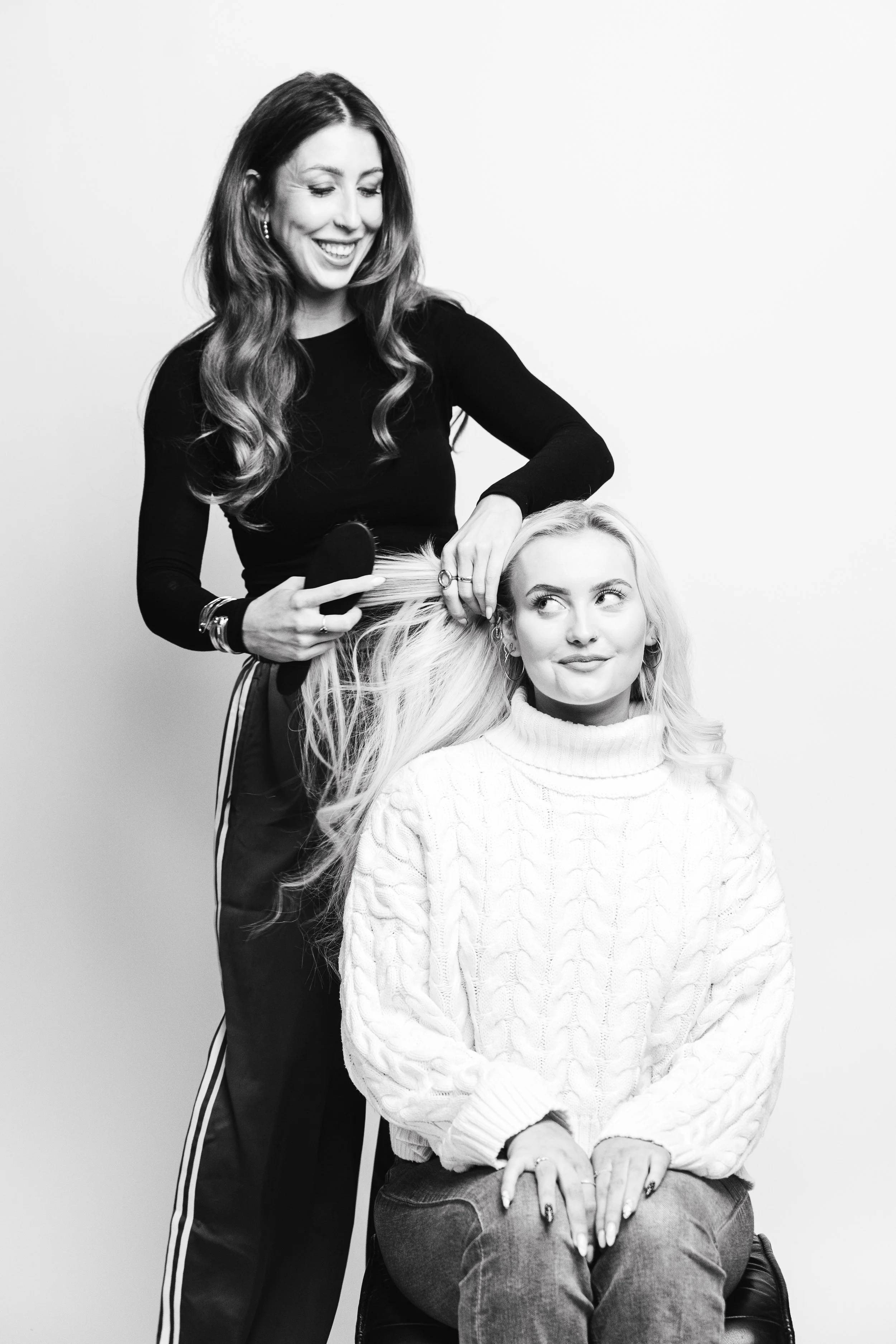 A woman with long wavy hair is styling the hair of a seated woman with long blonde hair and light skin using a hairbrush.
