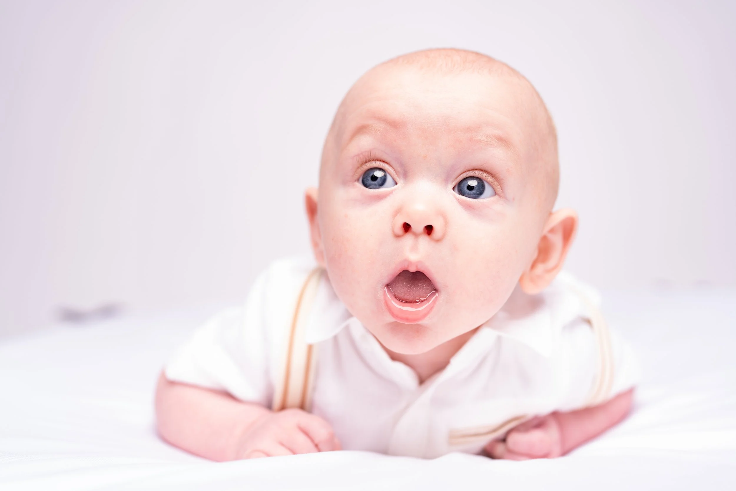 A baby with blue eyes and light skin, lying on a white surface, looking surprised or curious with mouth open.