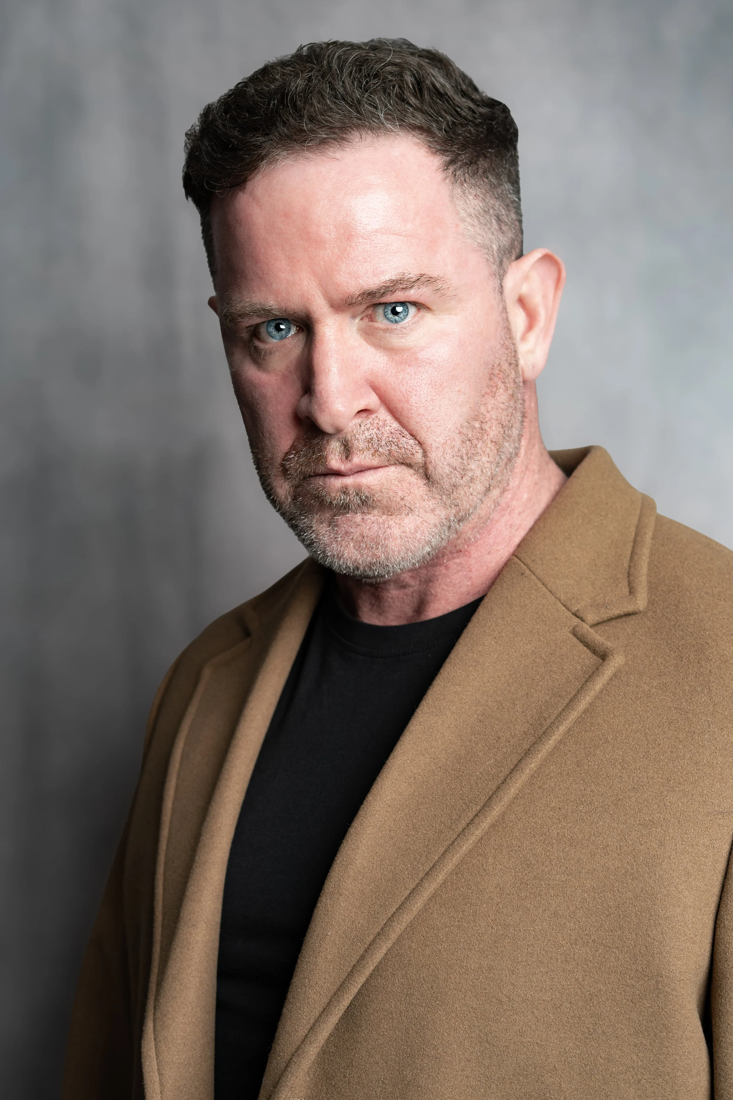 A middle-aged man with short, dark, curly hair and piercing blue eyes, wearing a light brown blazer over a black shirt, posing against a gray background.