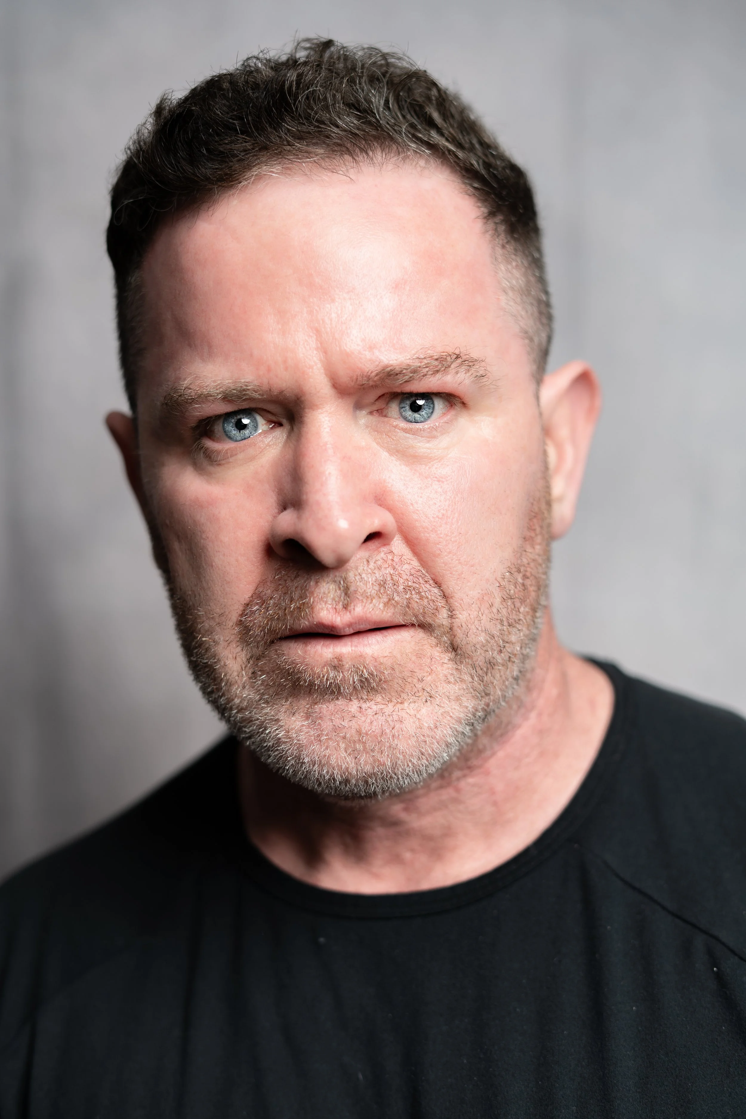 Close-up of a middle-aged man with short, dark hair, piercing blue eyes, and a beard, wearing a black shirt, looking directly at the camera against a neutral background.