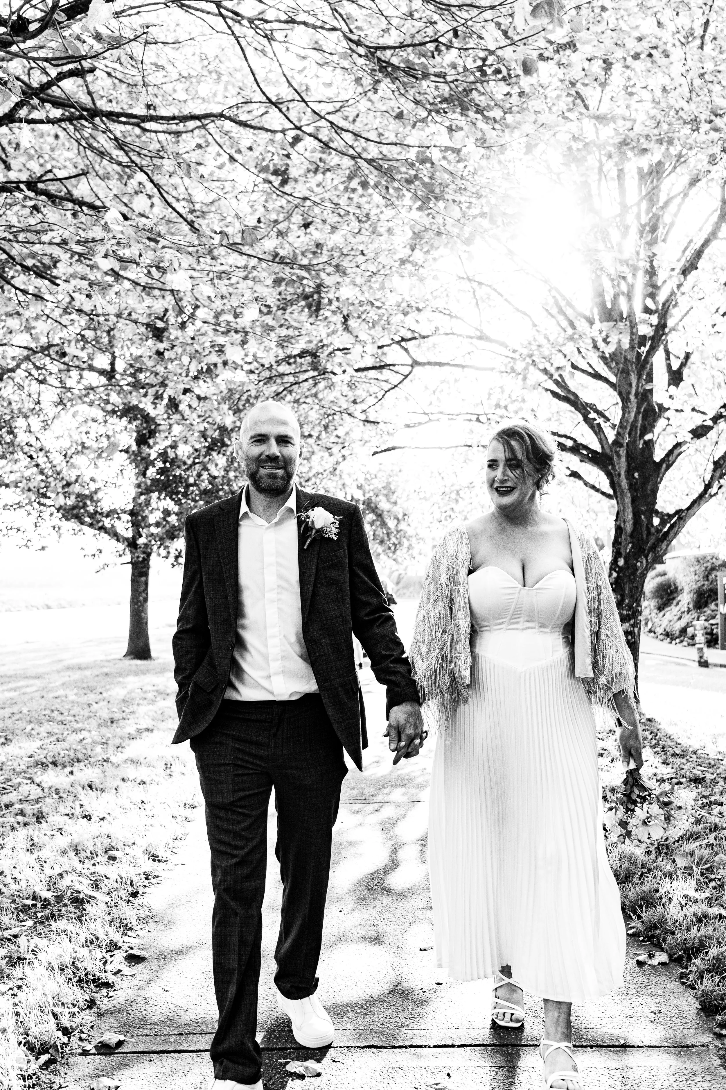 A black and white photo of a couple walking hand in hand outdoors, with trees and sunlight in the background. The woman is in a white dress, and the man is in a suit.