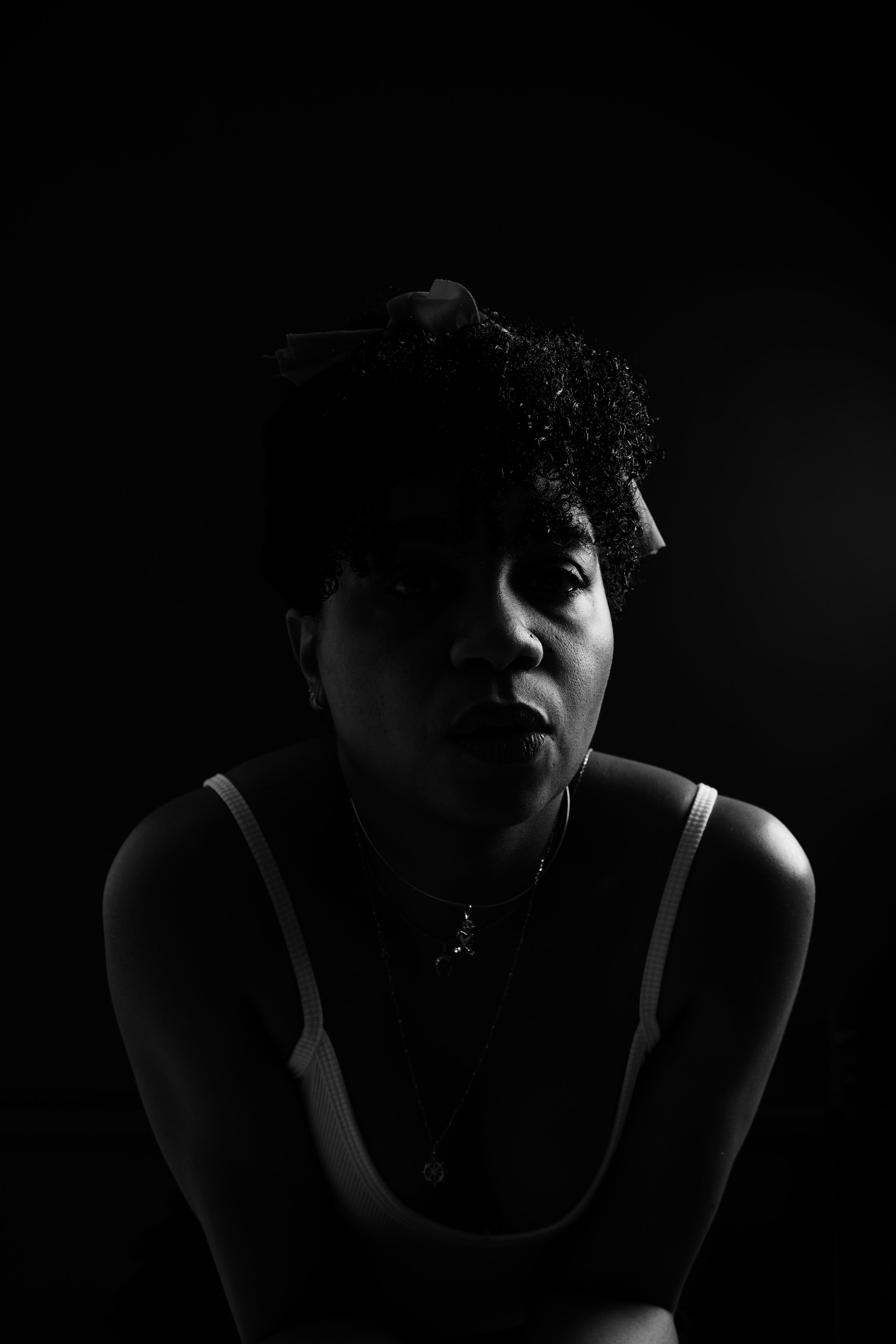 Black and white photo of a woman with short curly hair, wearing a tank top, with several necklaces, looking towards the camera with a serious expression.