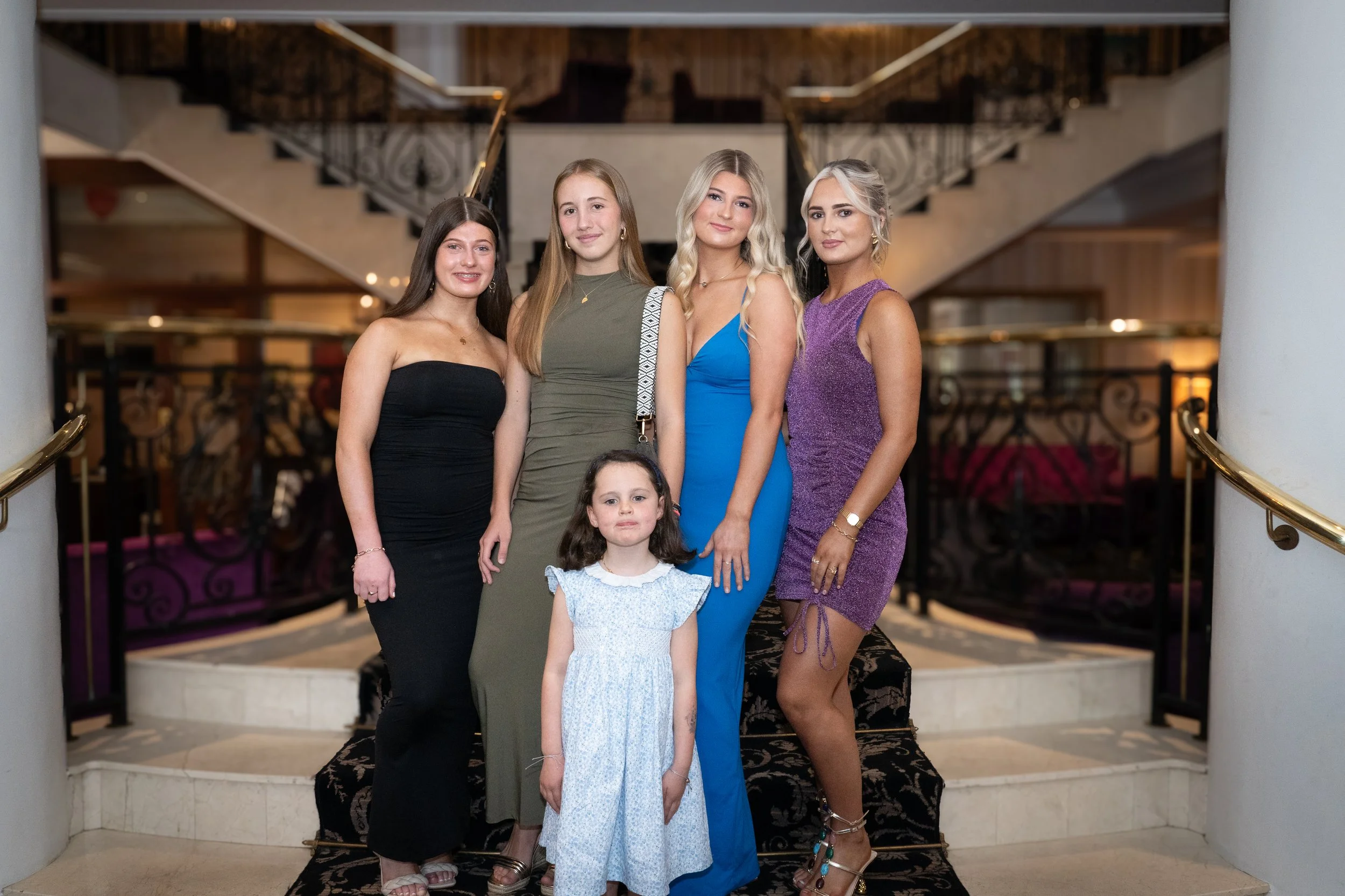 Group of six women and one young girl posing on a staircase in an elegant indoor setting.