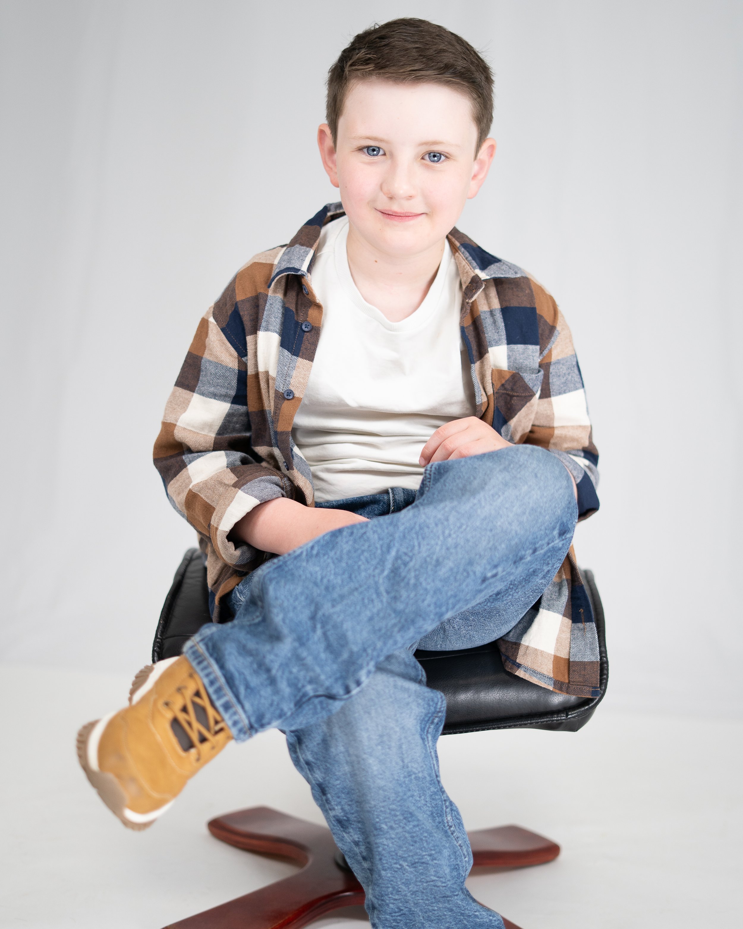 A young boy with short brown hair and blue eyes sitting on a black chair with one leg crossed over the other, wearing a white t-shirt, a brown and blue plaid shirt, blue jeans, and tan boots, smiling at the camera.