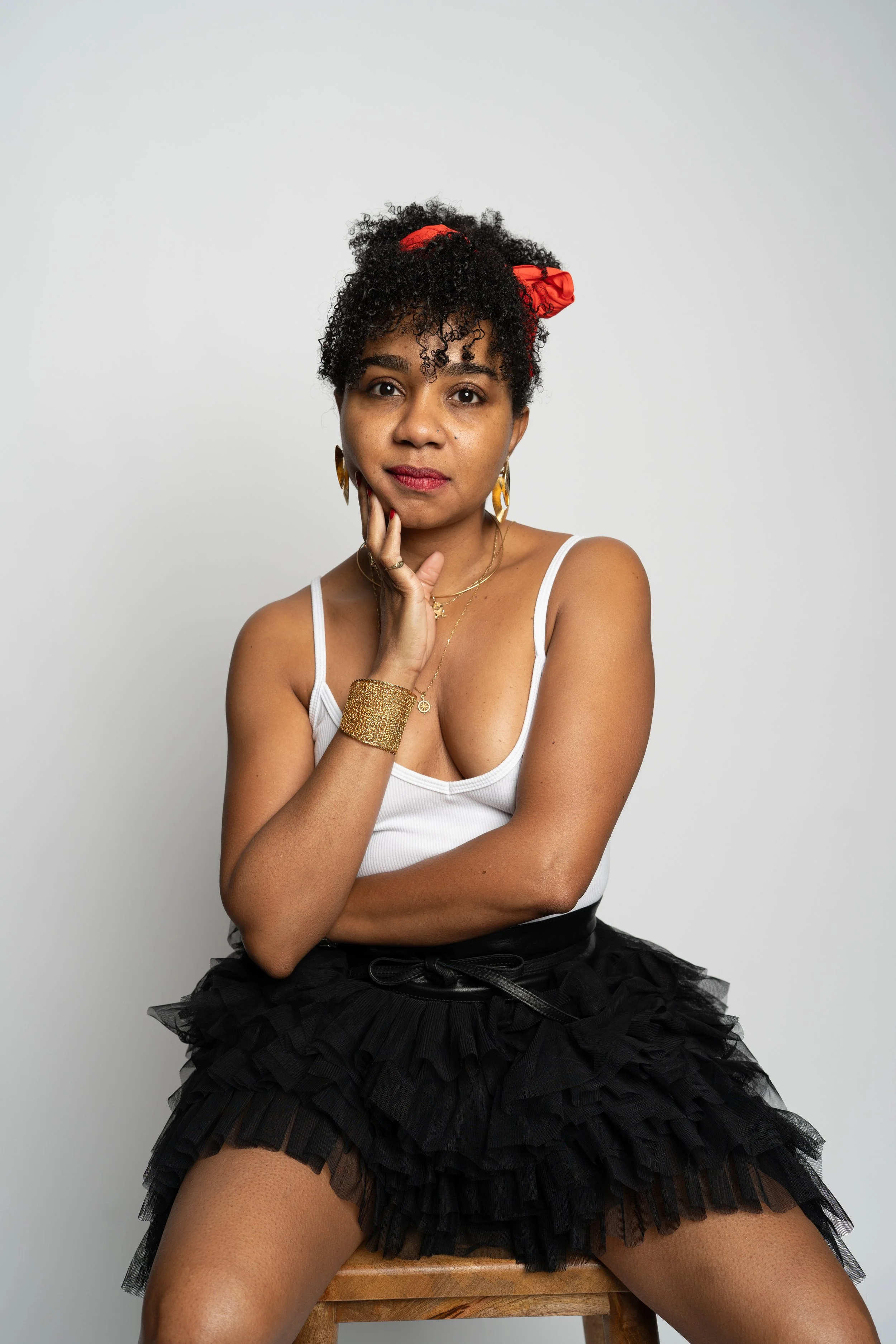 A woman with curly dark hair tied with a red ribbon, wearing a white tank top, a black ruffled skirt, and gold jewelry, posing seated on a wooden stool against a plain white background.