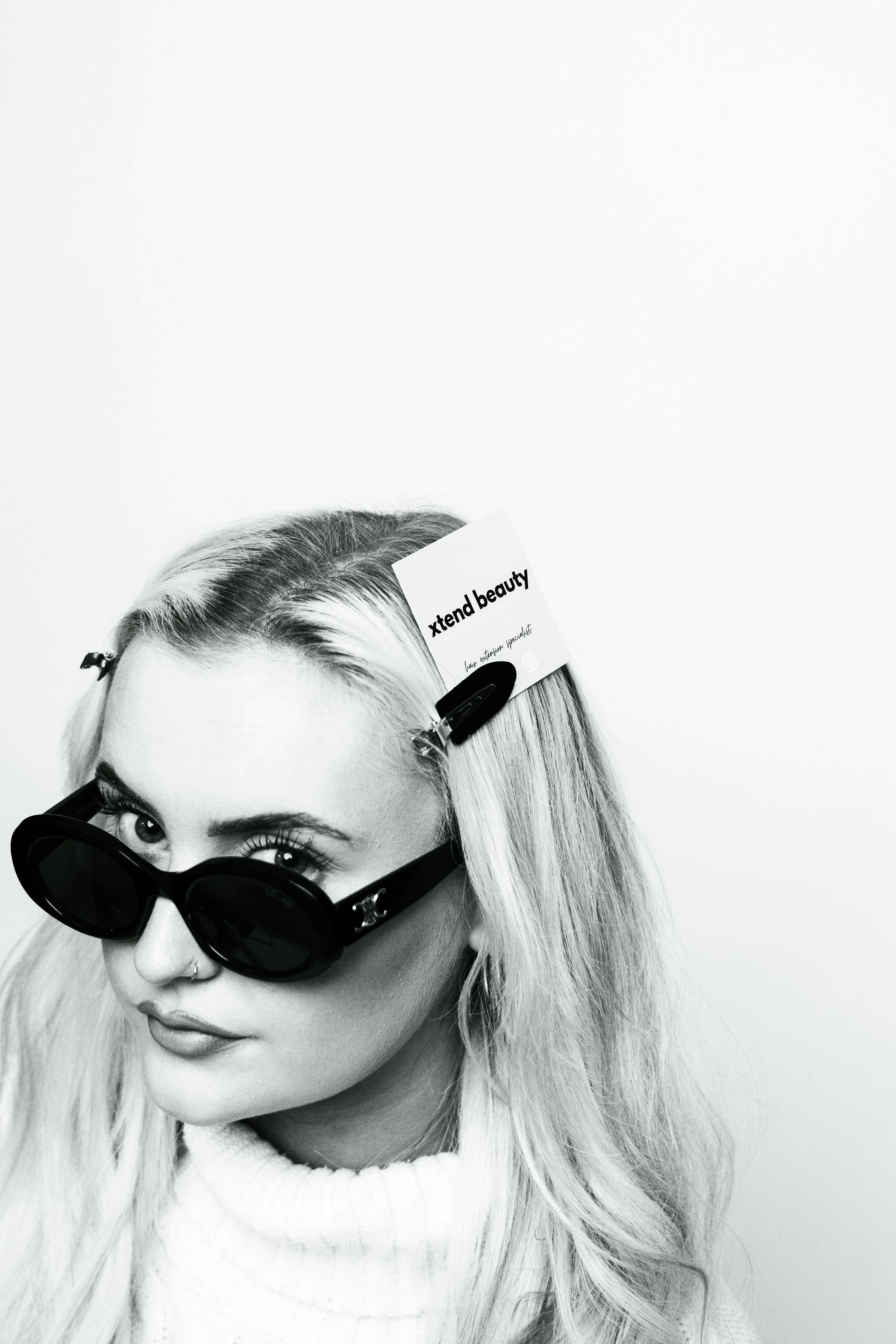 A woman with light-colored hair wearing dark sunglasses and a white turtleneck, with a tag clipped to her hair that reads 'xtend beauty'.