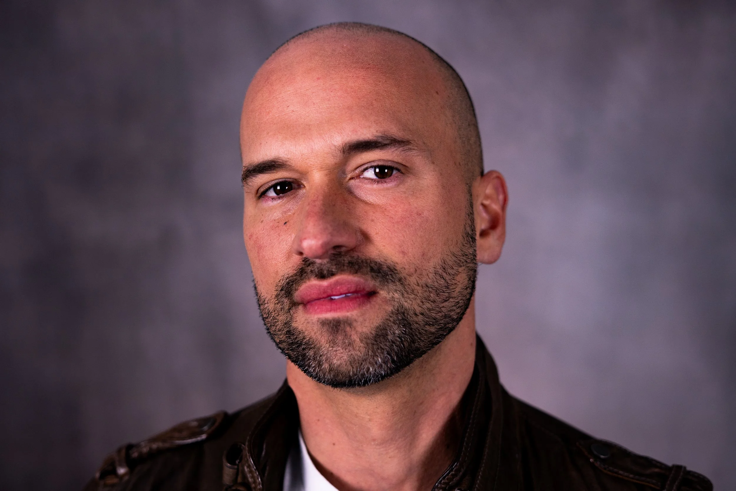 A close-up portrait of a bald man with a beard, wearing a leather jacket, against a plain gray background.