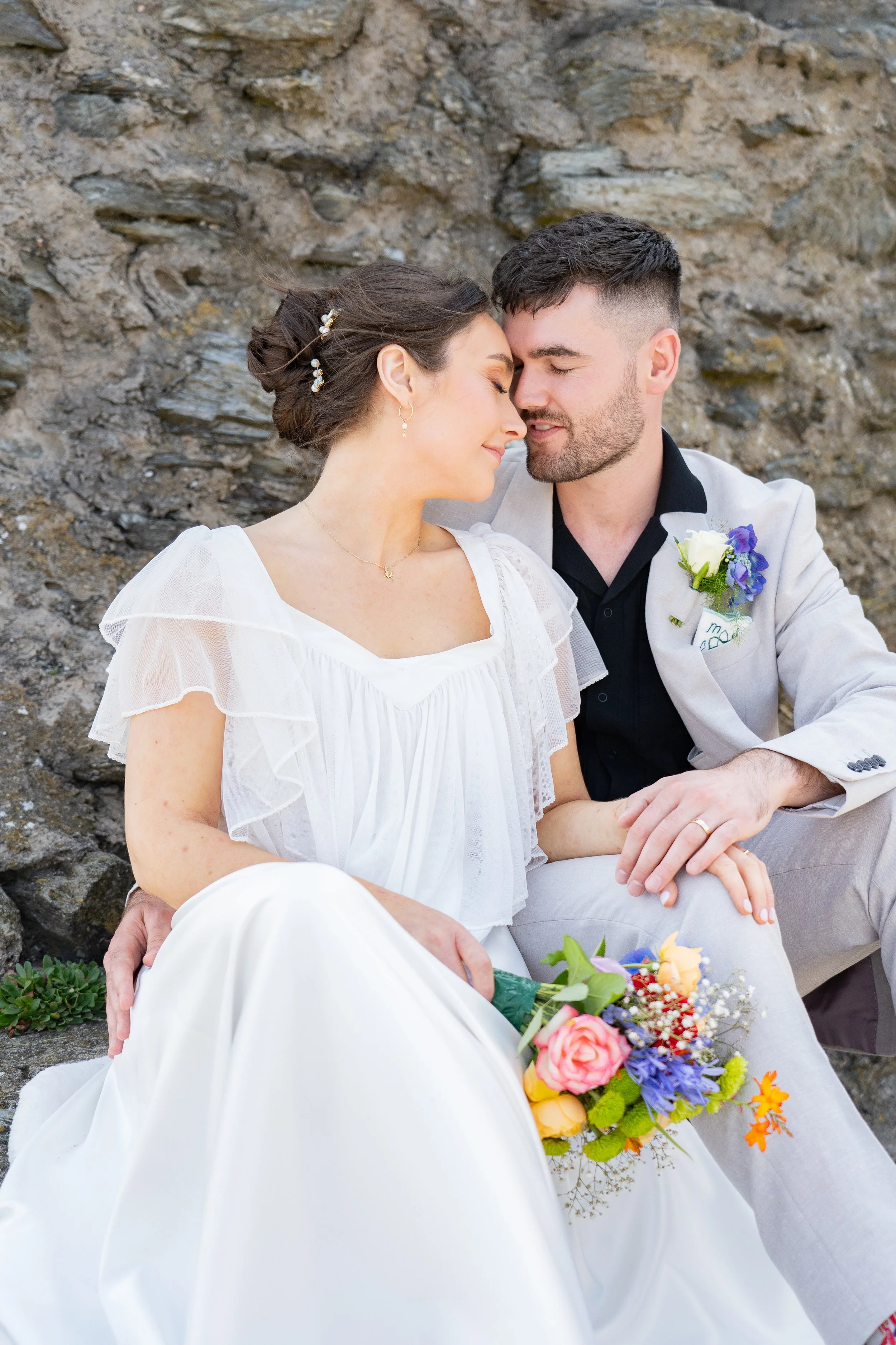 A bride and groom sitting close together, touching foreheads with eyes closed, against a rocky background. The bride holds a colorful bouquet of flowers, and the groom wears a light suit with a boutonnière.
