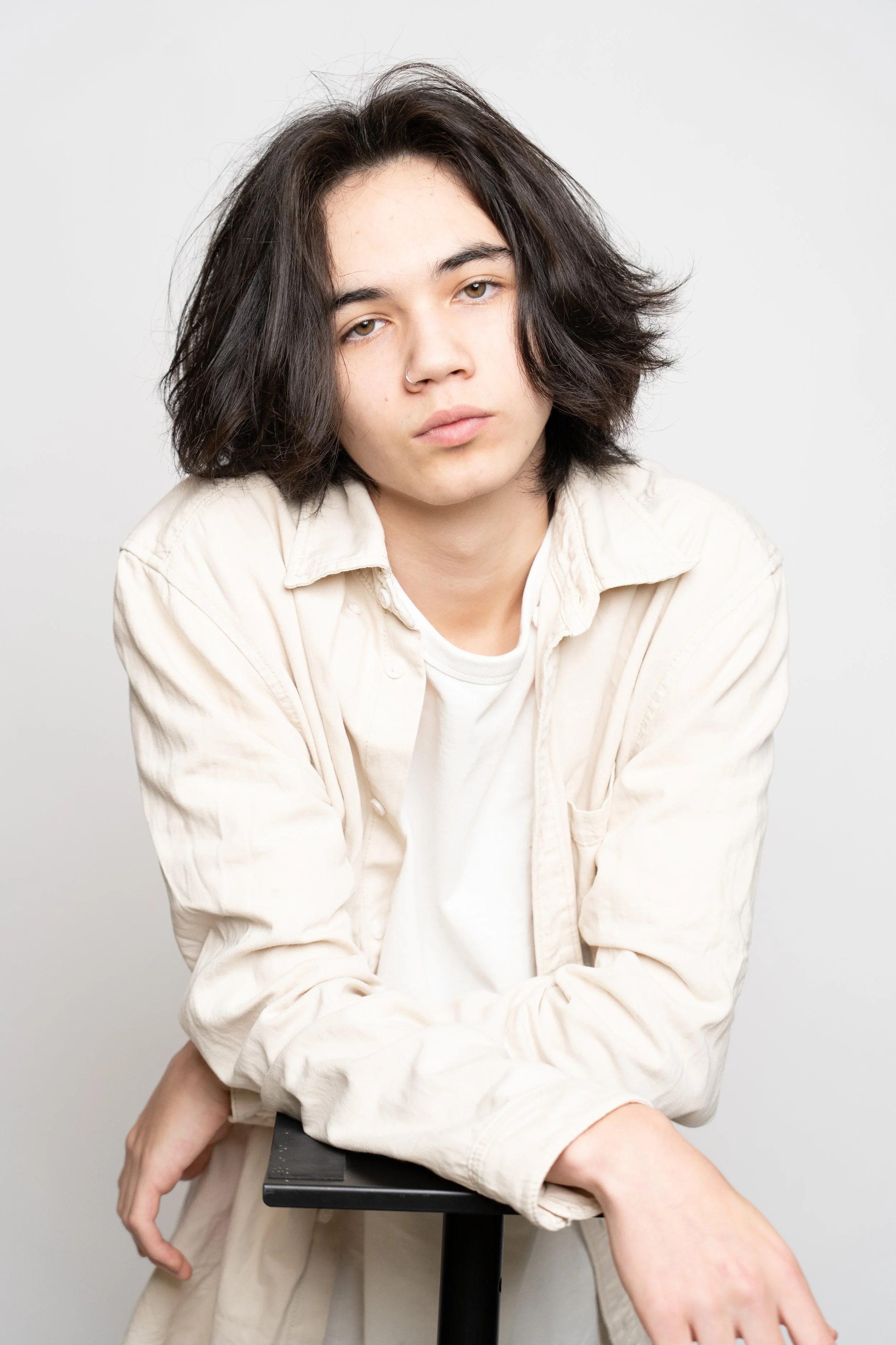 A young person with medium-length dark hair, wearing a light-colored shirt and t-shirt, leaning on a black stand, looking directly at the camera with a neutral expression.