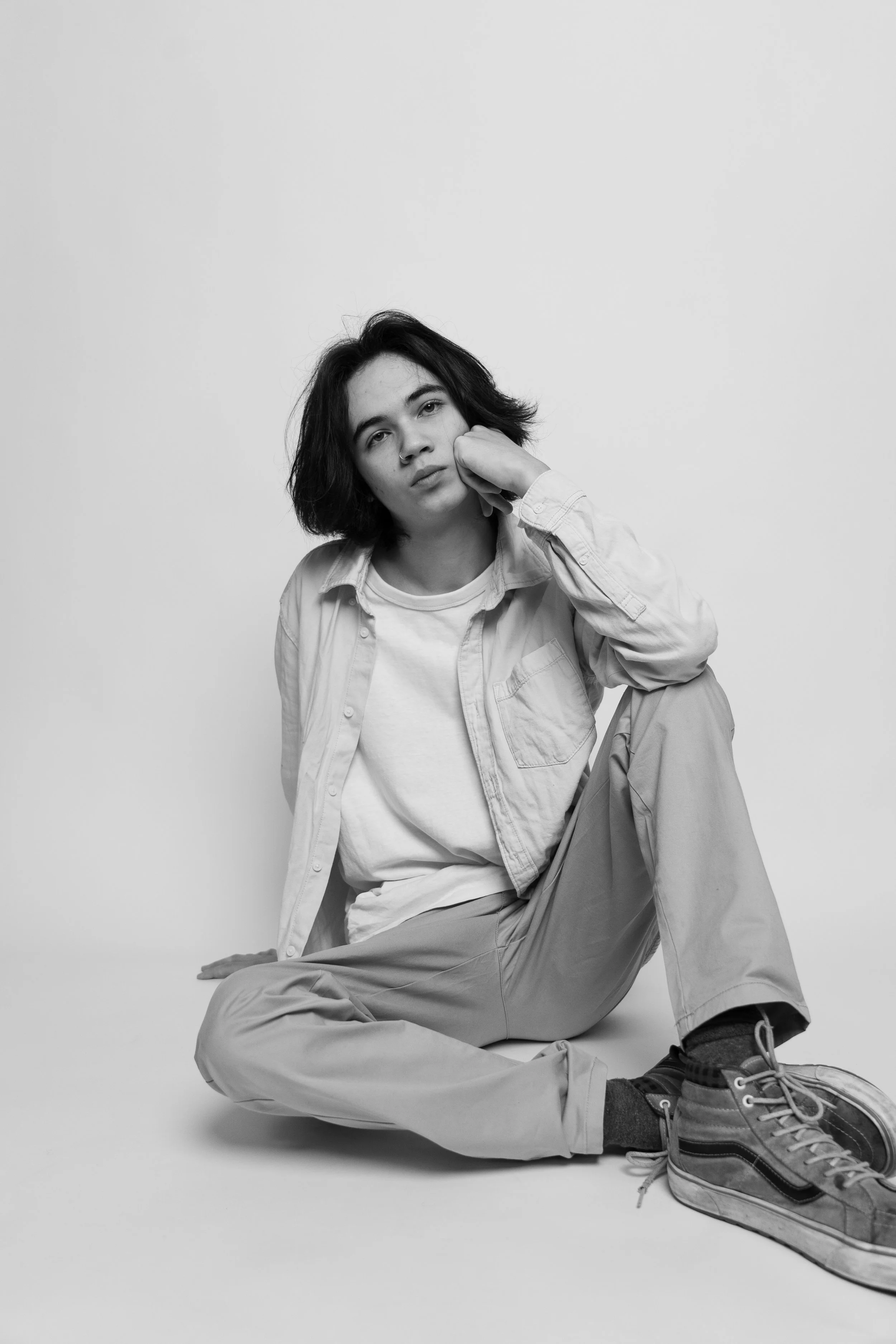 Black and white photo of a young woman sitting on the floor with her legs bent, resting her left hand on her face, wearing a casual collared shirt, T-shirt, loose pants, and sneakers, against a plain background.