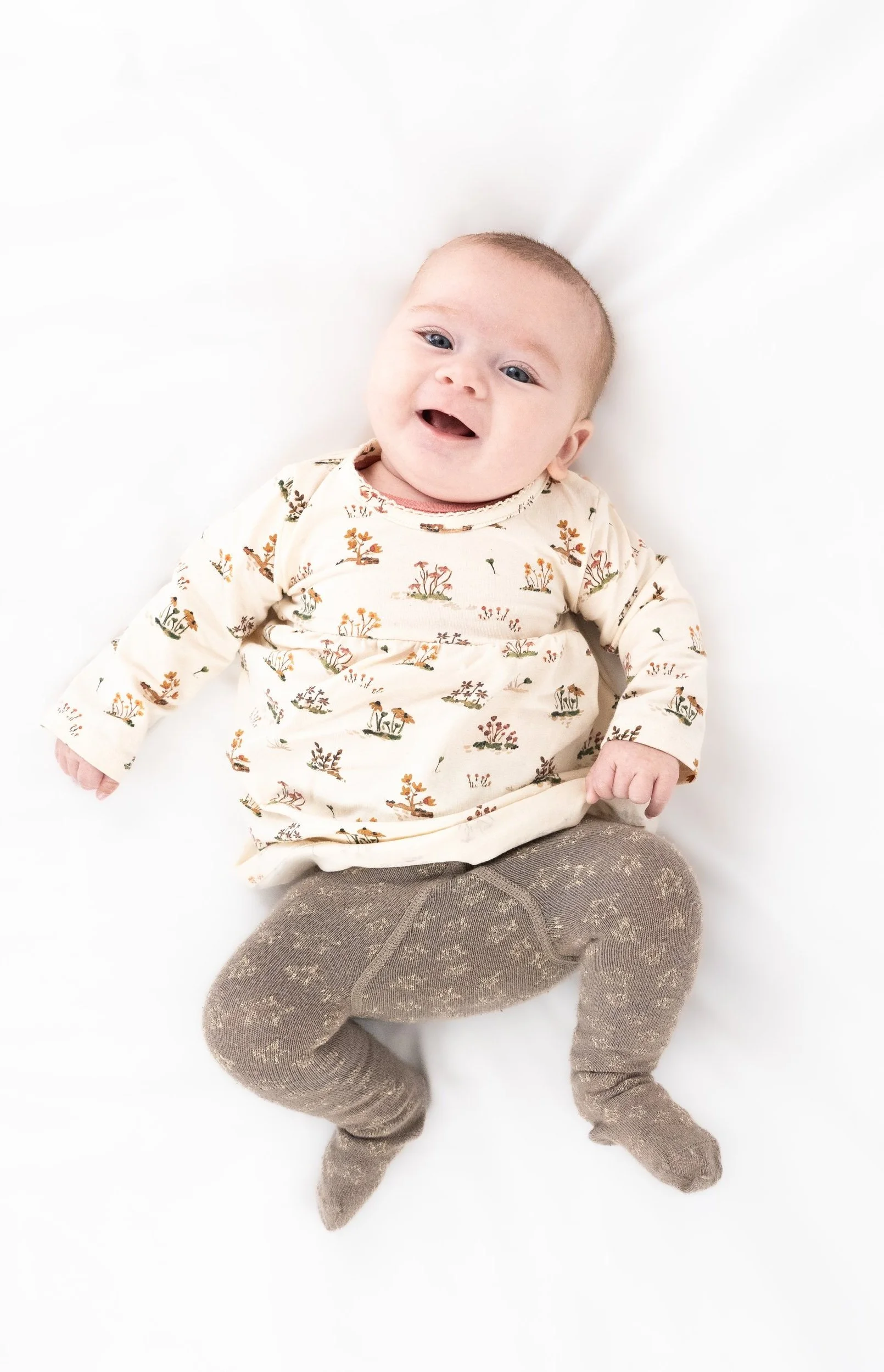 A smiling baby lying on a white surface, wearing a floral long-sleeve shirt and beige patterned tights.