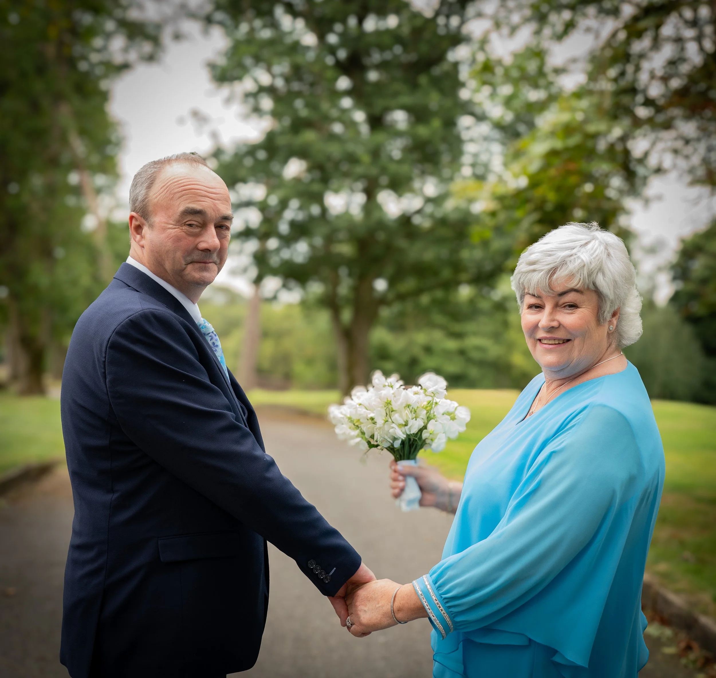 An older man and woman standing outdoors on a pathway surrounded by trees, holding hands. The woman is smiling and holding a bouquet of white flowers, while the man has a neutral expression. The woman is wearing a light blue dress, and the man is wea