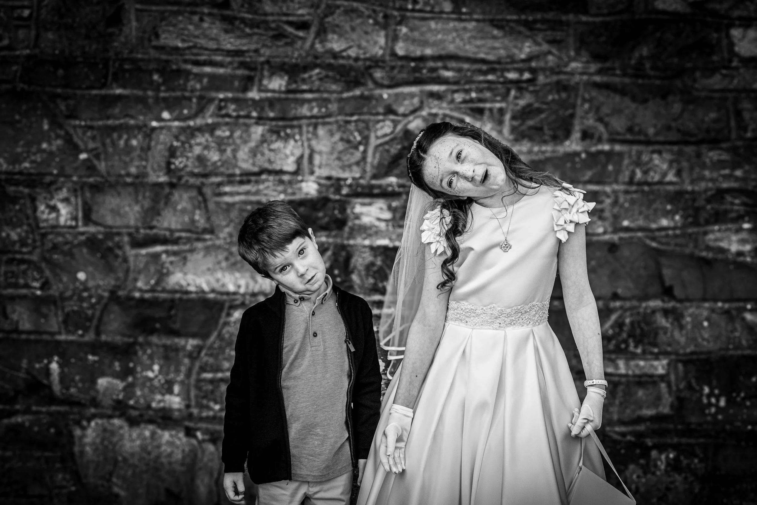 A girl in a wedding dress with a veil and gloves, and a boy in casual clothing, both tilting their heads to the side, standing against a stone wall background.