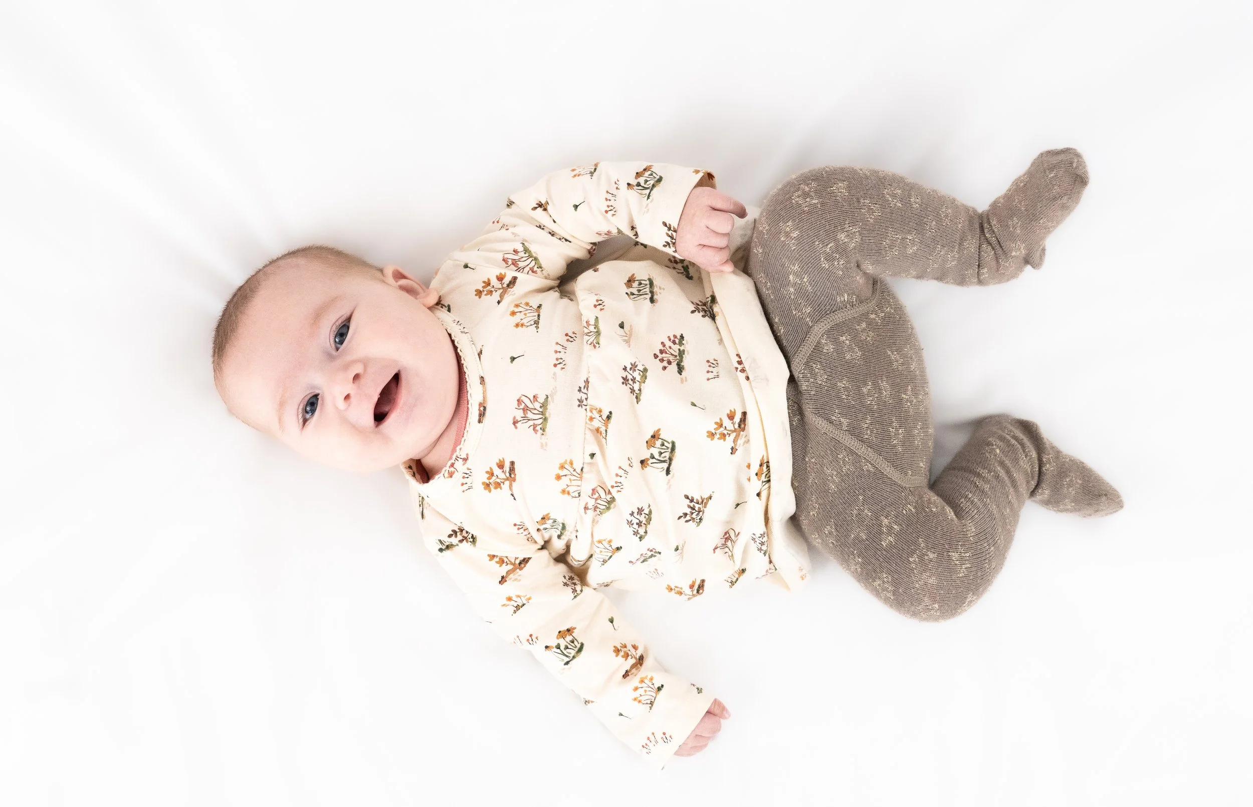 Baby lying on white surface, smiling, wearing a long-sleeved onesie with a nature print and gray knitted leggings.