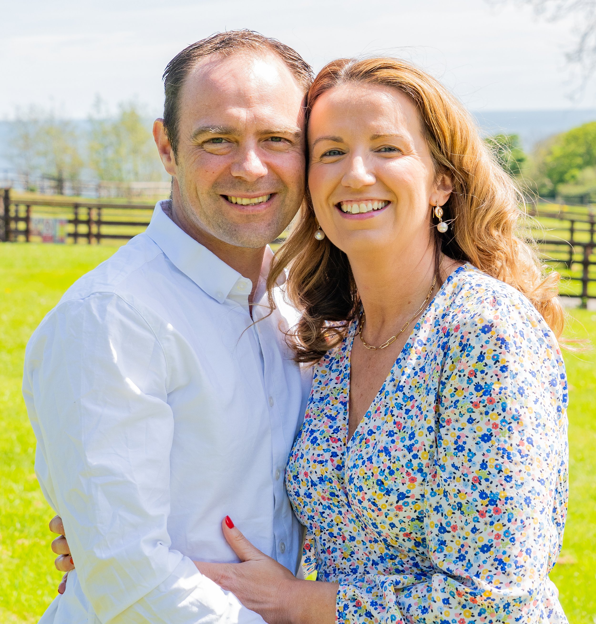 A smiling couple outdoors in a sunny field, embracing each other with a wooden fence and greenery in the background.