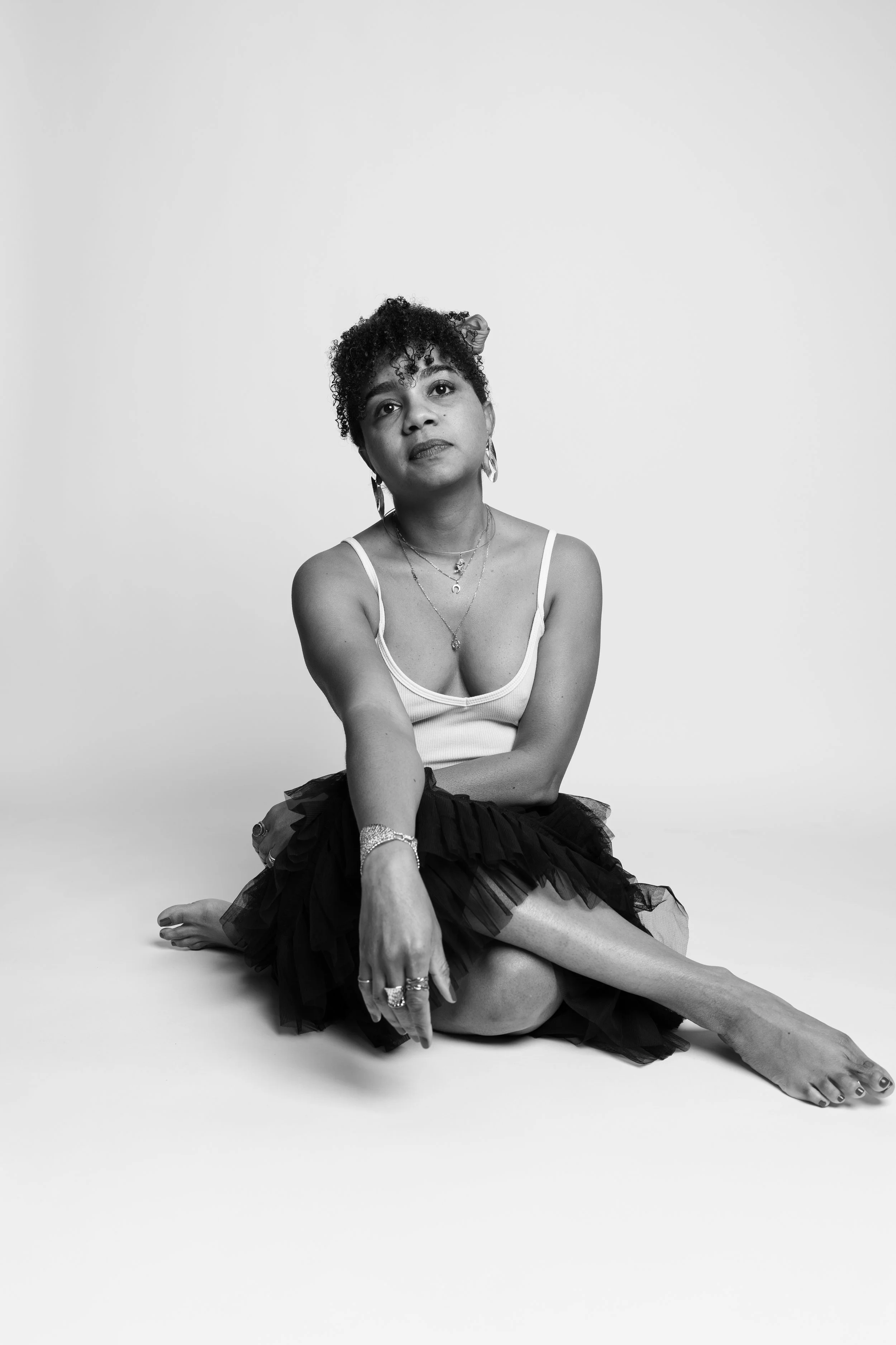 Black and white portrait of a woman with curly hair, sitting barefoot on the floor with a plain white background, wearing a tank top and a ruffled skirt, accessorized with jewelry.
