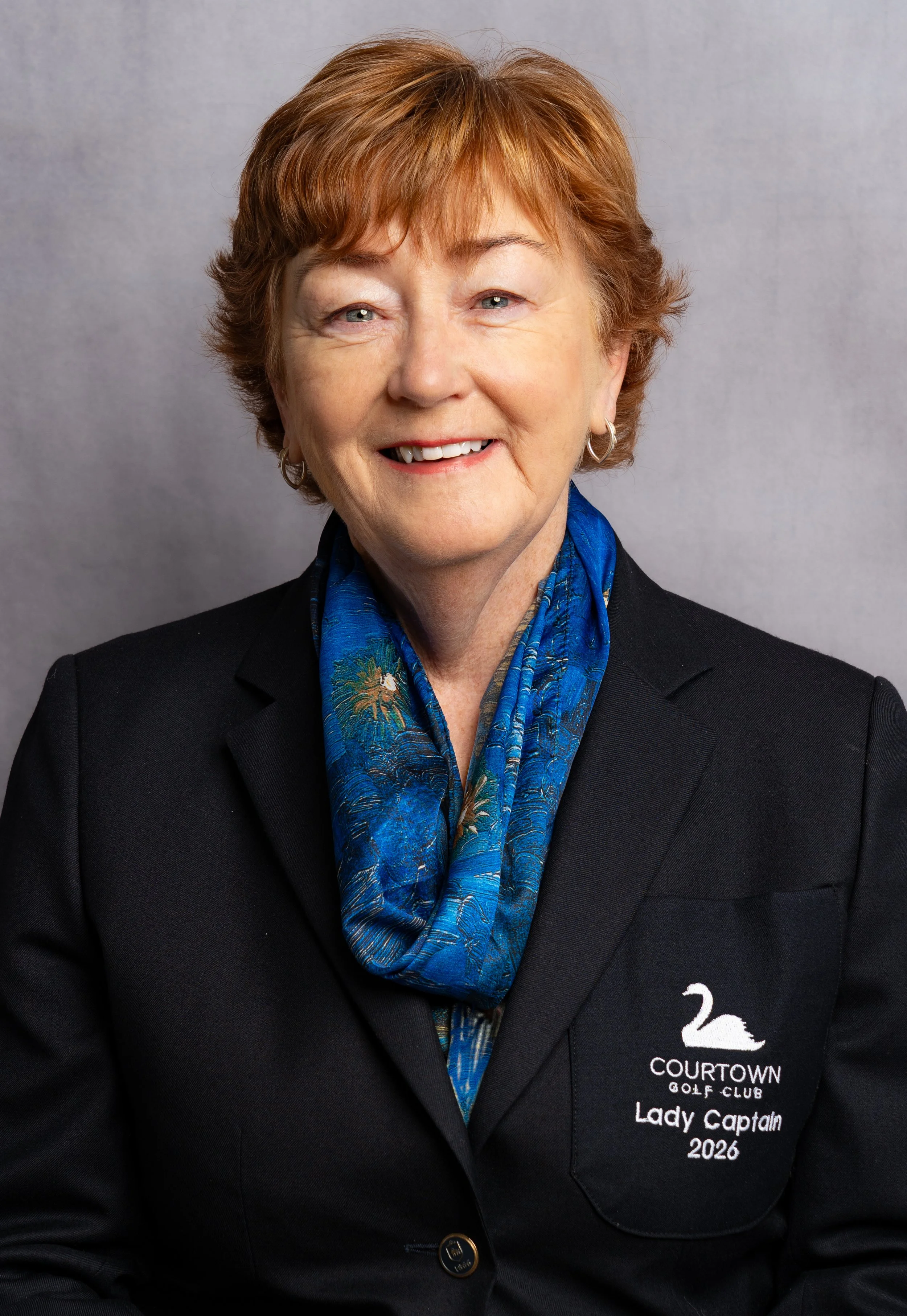 An older woman with short, red hair, smiling, wearing a black blazer with a 'Lady Captain 2026' embroidered on the pocket, a blue patterned scarf, and earrings, standing against a gray background.