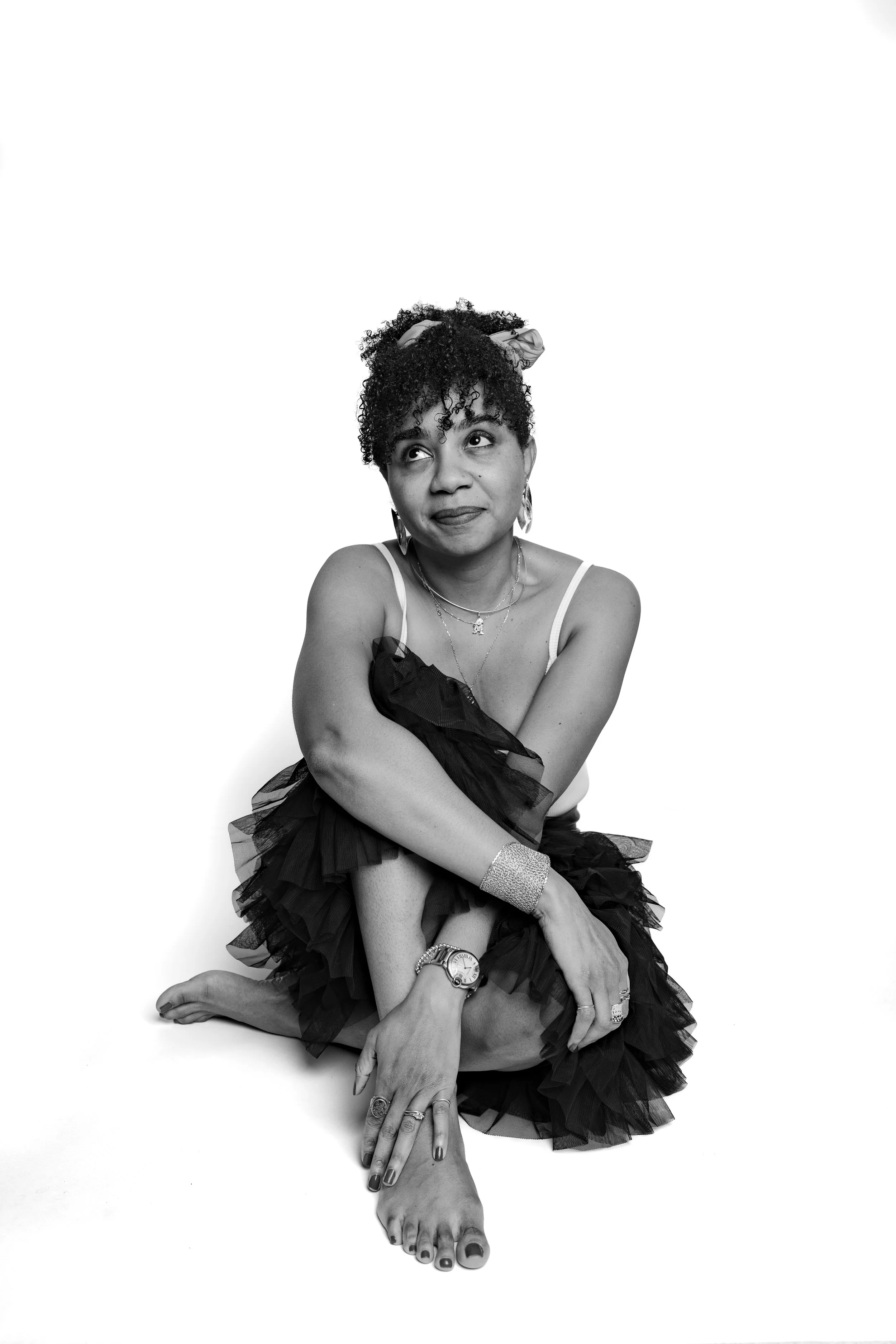 Black and white photo of a woman with curly hair, sitting on the floor with cross-legged pose, wearing jewelry and a ruffled skirt, looking thoughtfully upward.