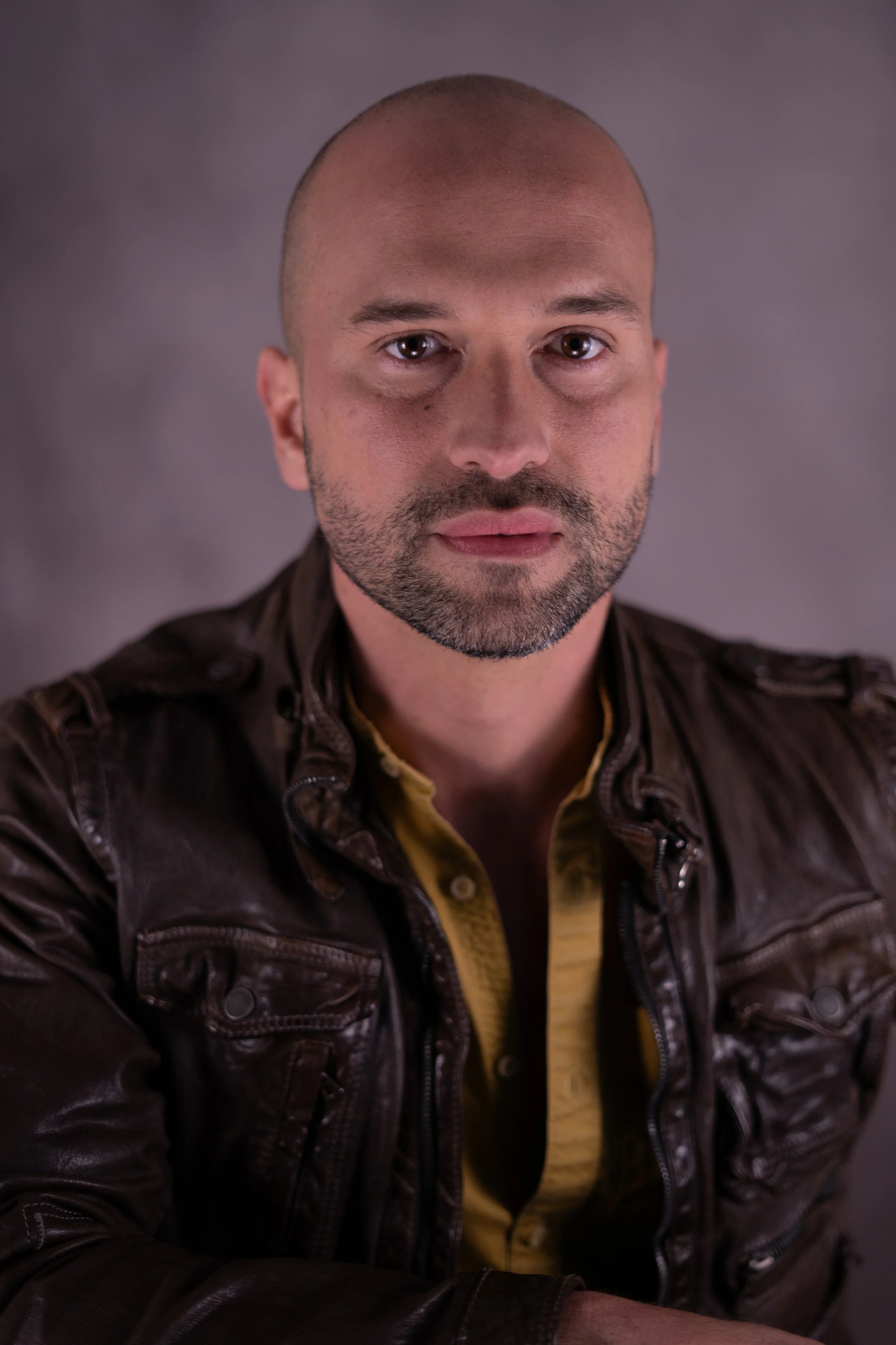 A man with a shaved head, beard, and mustache wearing a brown leather jacket and a yellow shirt, looking directly at the camera against a gray background.