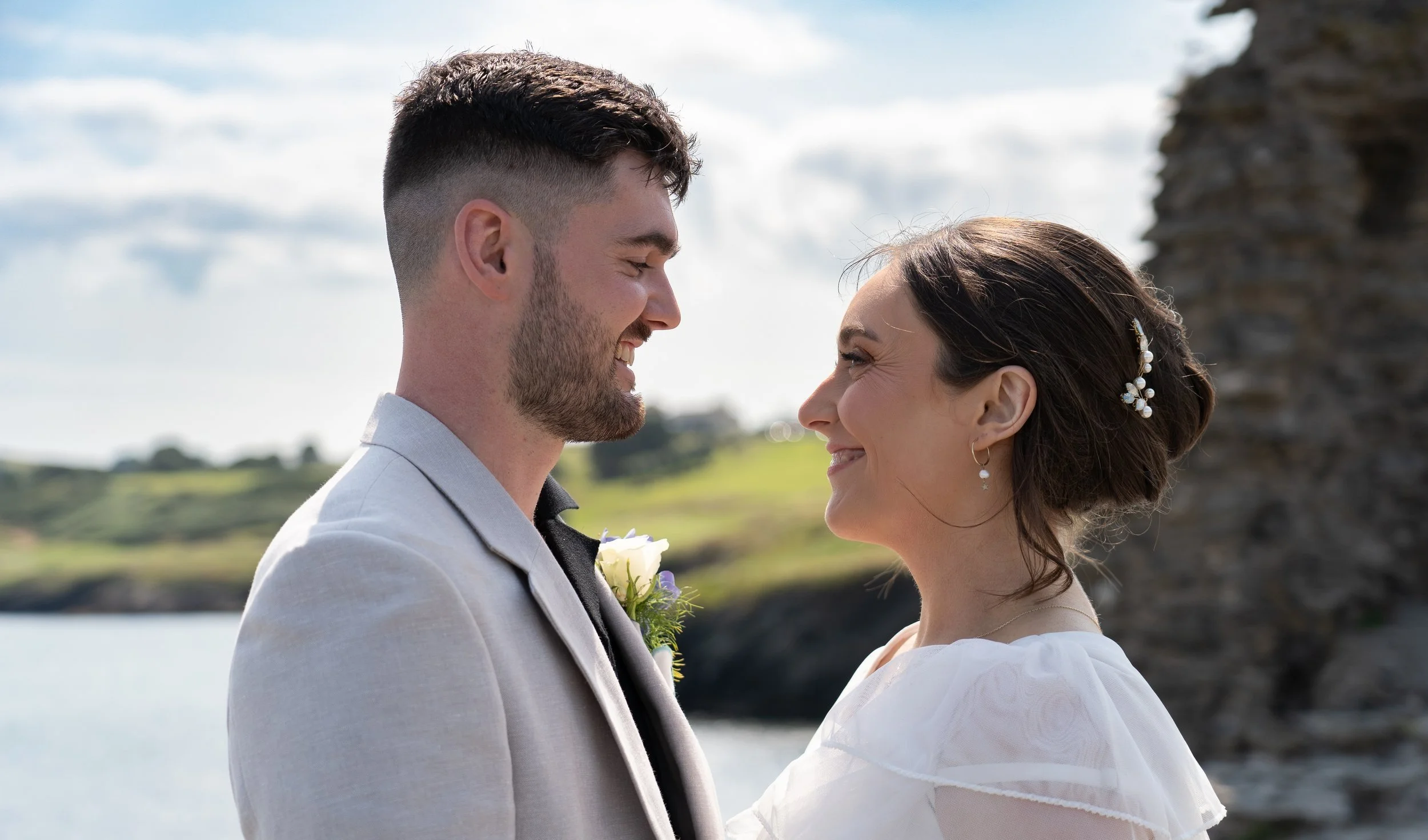 A bride and groom smiling and gazing into each other's eyes outdoors near water and rocky cliffs.