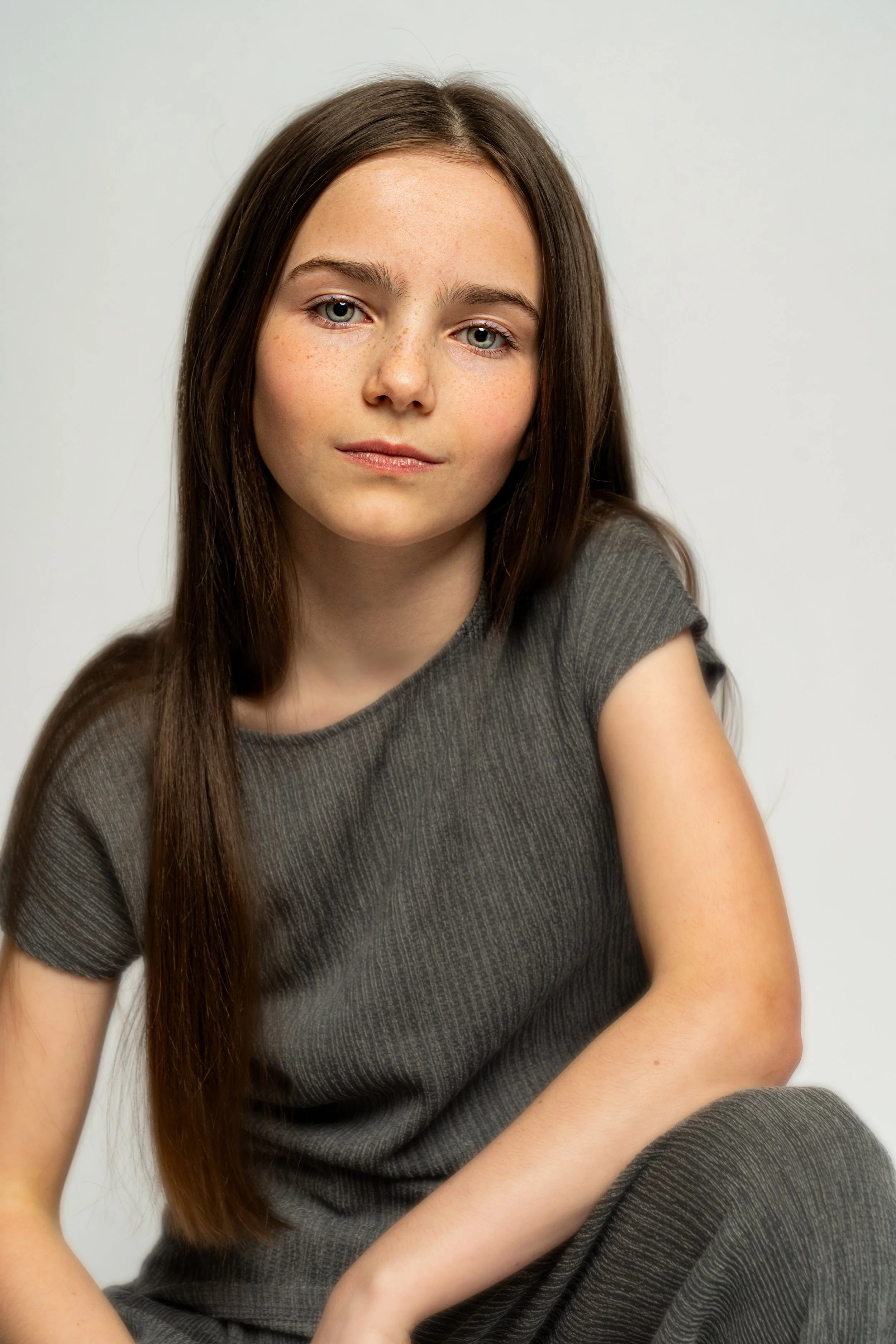 A young girl with long brown hair, light skin, and freckles, wearing a gray ribbed shirt, sitting against a plain light background, looking at the camera.