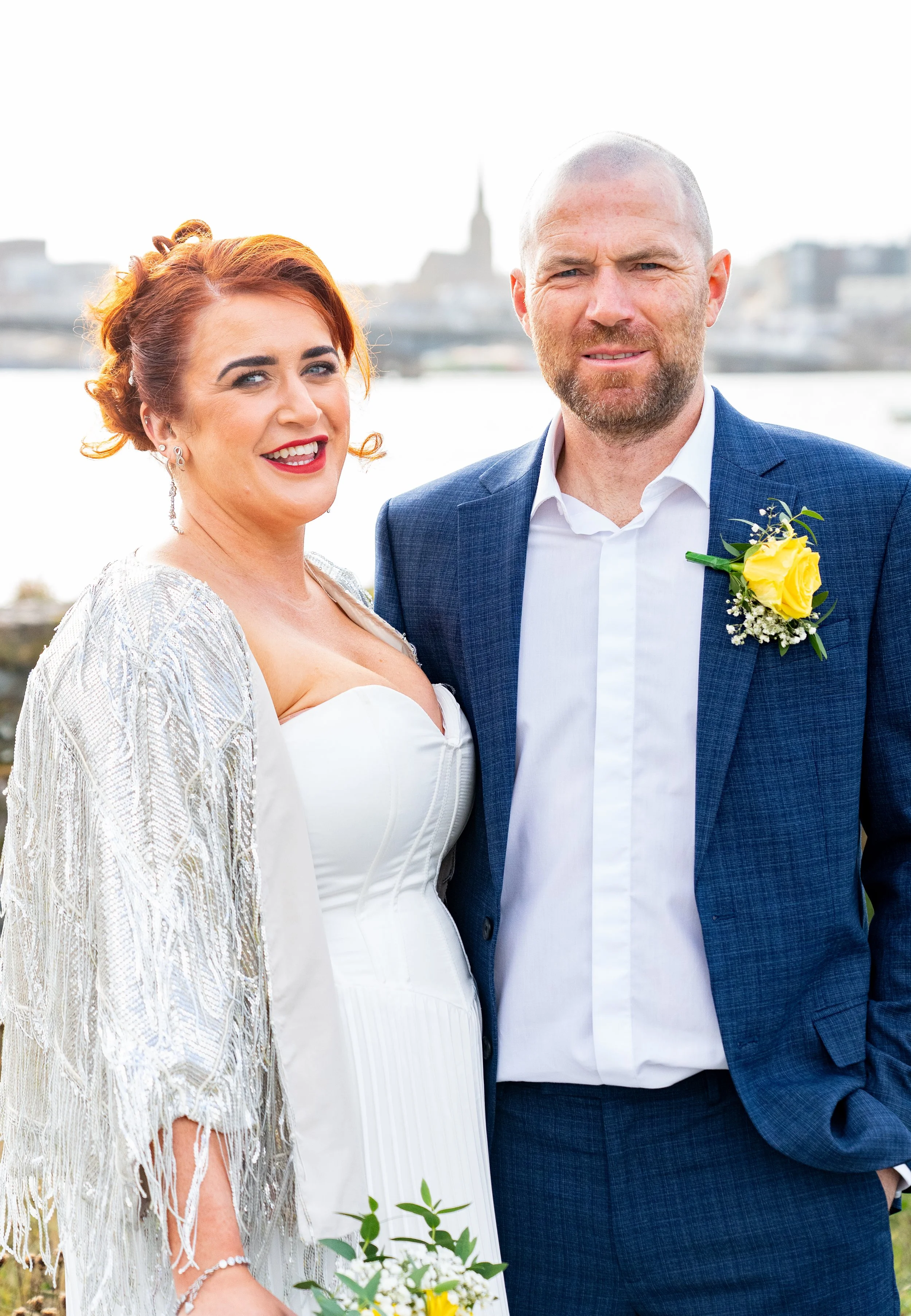 A newlywed couple standing outdoors near a body of water with a cityscape and church spire in the background. The woman has red hair, wearing a white wedding dress and silver shawl, holding a small bouquet. The man has a beard, wearing a blue suit wi