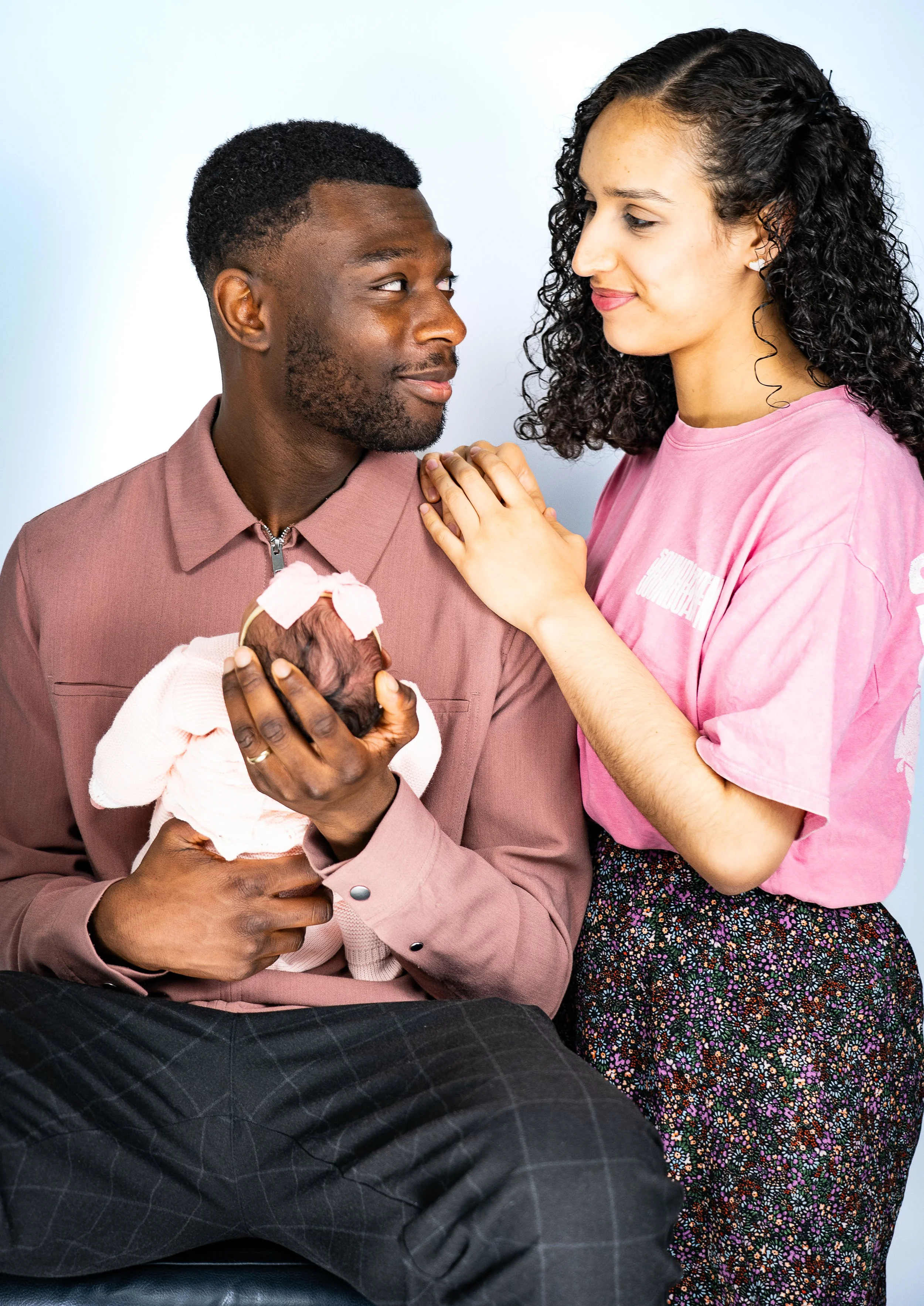 A man holding a newborn baby in his arms, sitting and looking at a woman who is touching his shoulder, in a hospital or home setting.